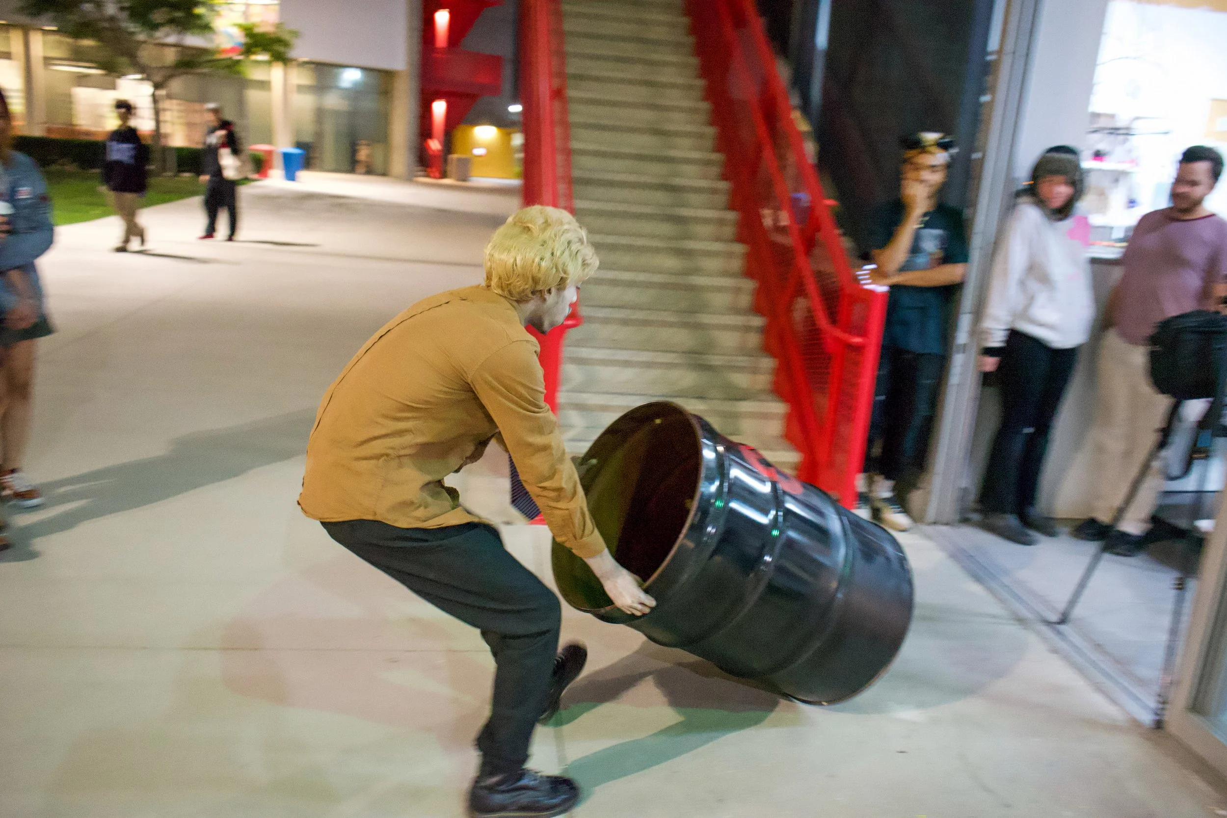 A man with blonde hair wearing a tan jacket and black pants is pushing a black barrel at night outside a building, with a group of people watching from inside and outside.