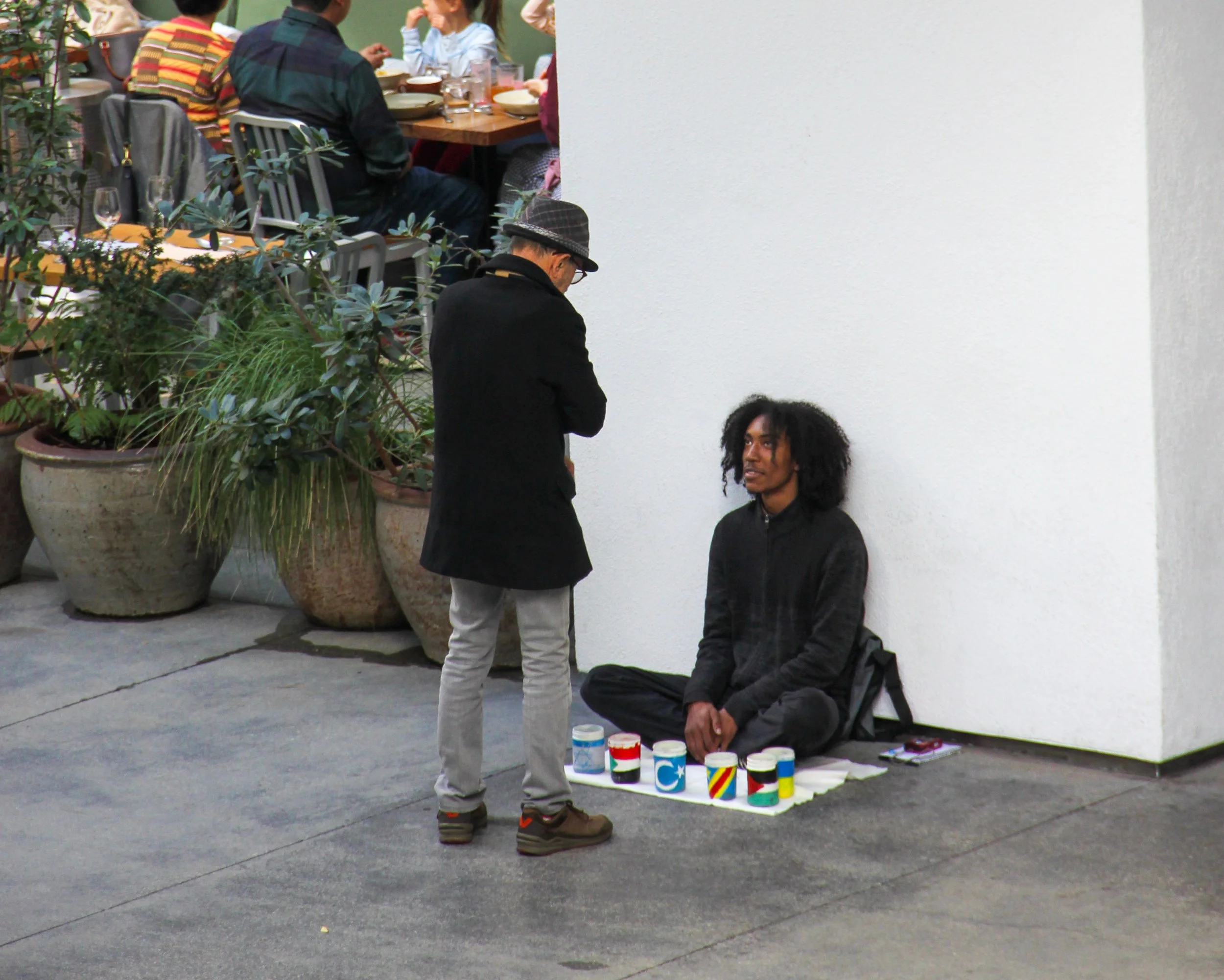 A man with long curly hair sits on the sidewalk against a white wall, with various colorful cups or cans arranged in front of him on a mat. A person in a black coat and gray pants stands nearby, talking to him. In the background, a group of people sit at a table in a restaurant.