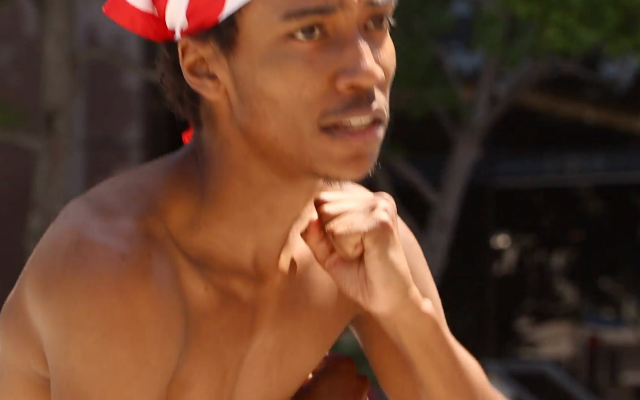 Close-up of a shirtless man wearing a red and white bandana, sitting outdoors at night with a thoughtful expression.
