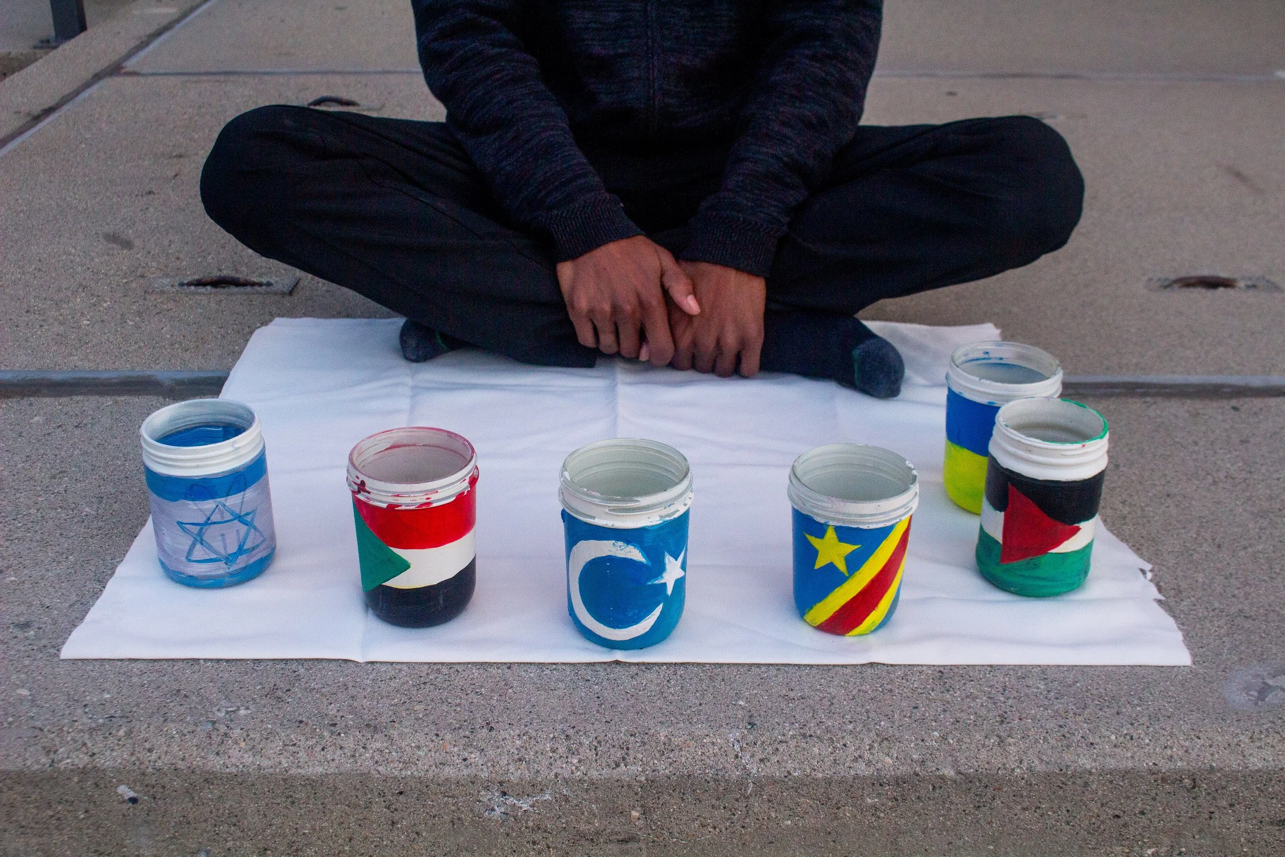 A person sitting cross-legged on a sidewalk with five painted jars in front of them, each decorated with the flag of a different country, on a white cloth.