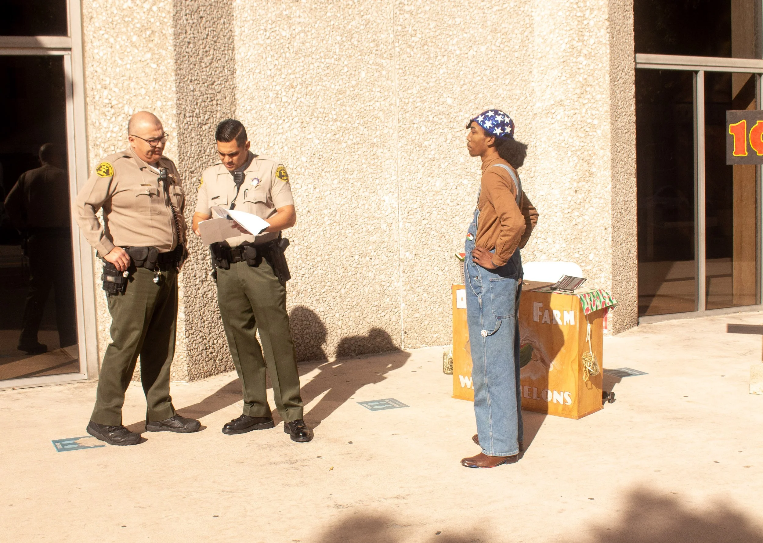 Two police officers standing and reviewing documents, a woman standing with hands on hips near a stand with a sign reading "Watermelons", outside a building with glass doors.