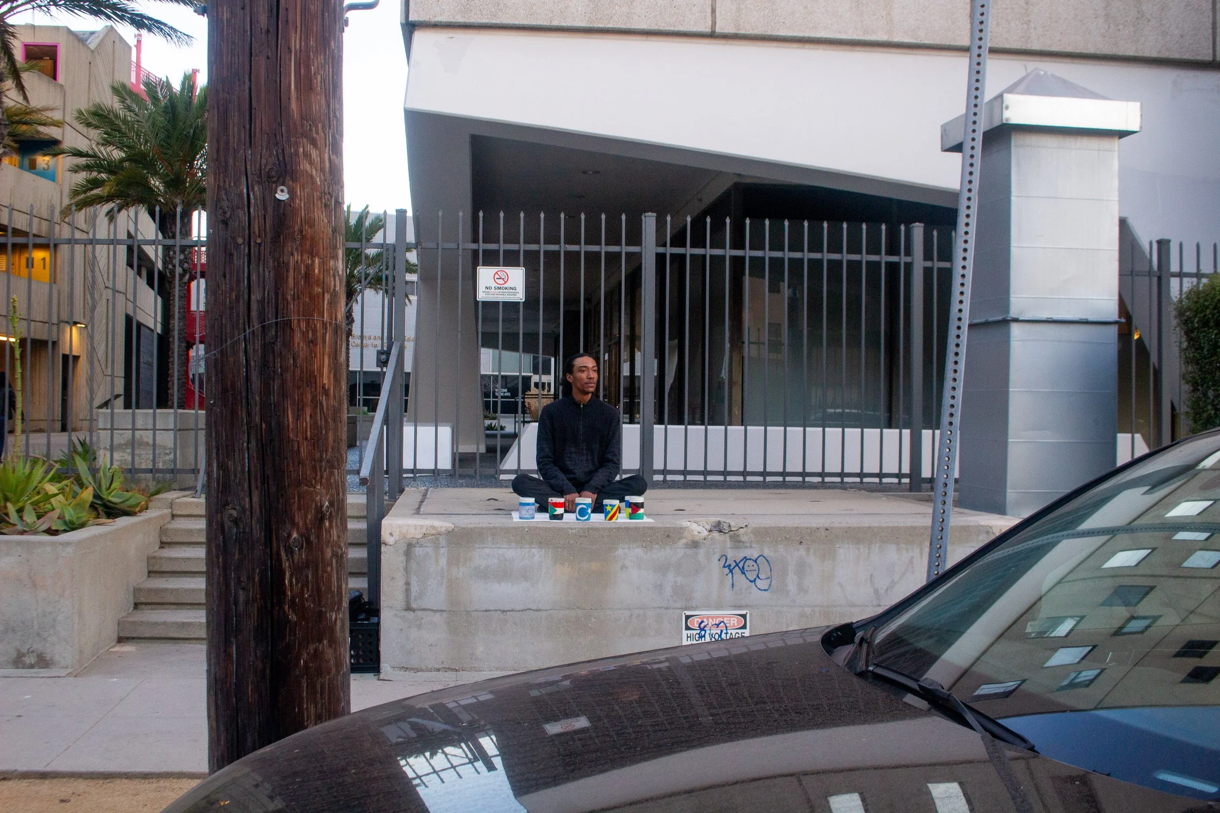 A man sitting on a concrete ledge with several painted cups in front of him on a city sidewalk, adjacent to a building with a metal fence and stairs.