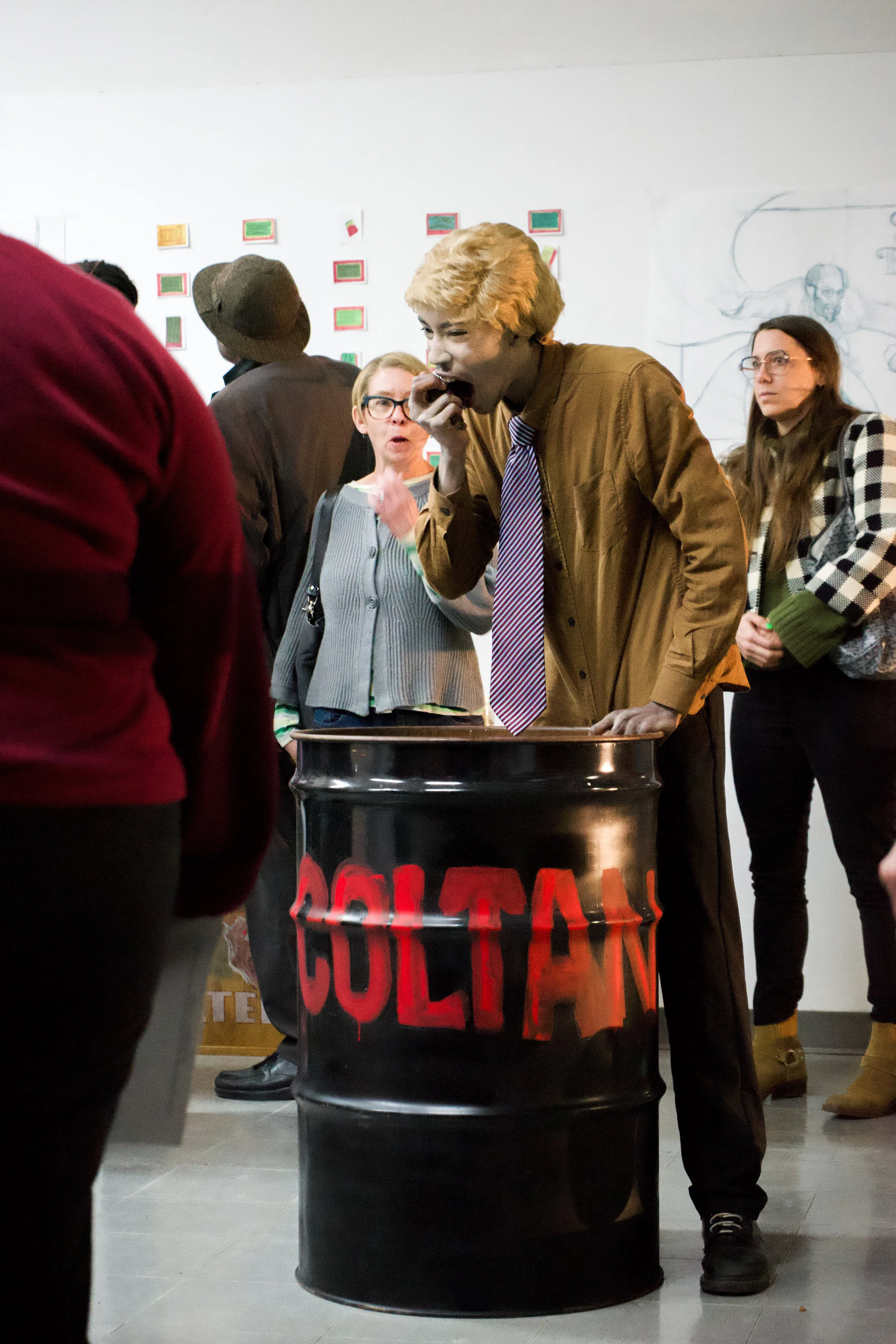 A man with blonde hair wearing a brown jacket and a striped tie, standing inside a black barrel with the word 'CULTAIN' spray painted in red, eating a snack. Several people around him are watching, with varied expressions. The scene appears to be in 