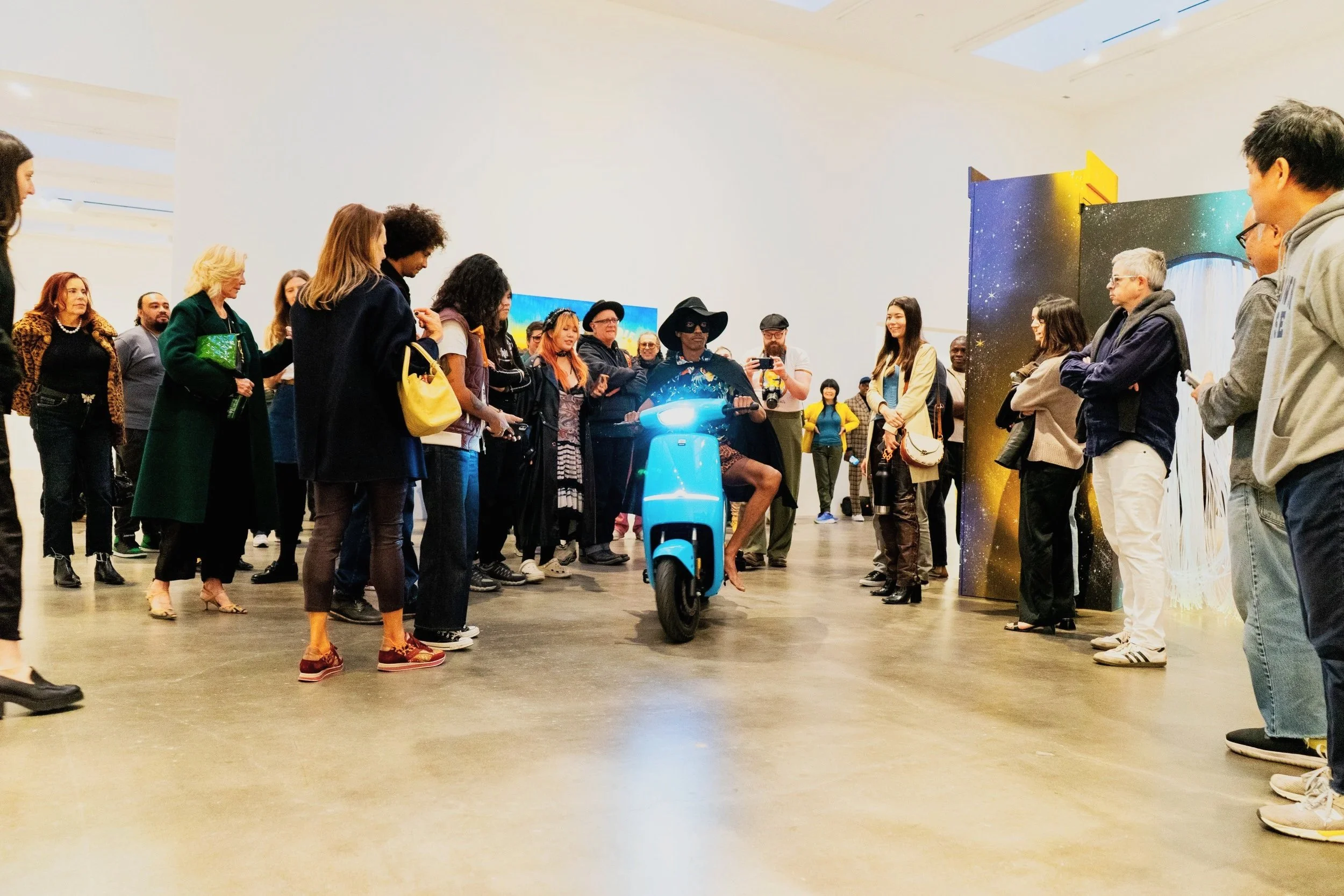 A woman in a black dress and sunglasses riding a bright blue scooter through a crowd at an art gallery or event, surrounded by people taking photos and watching.