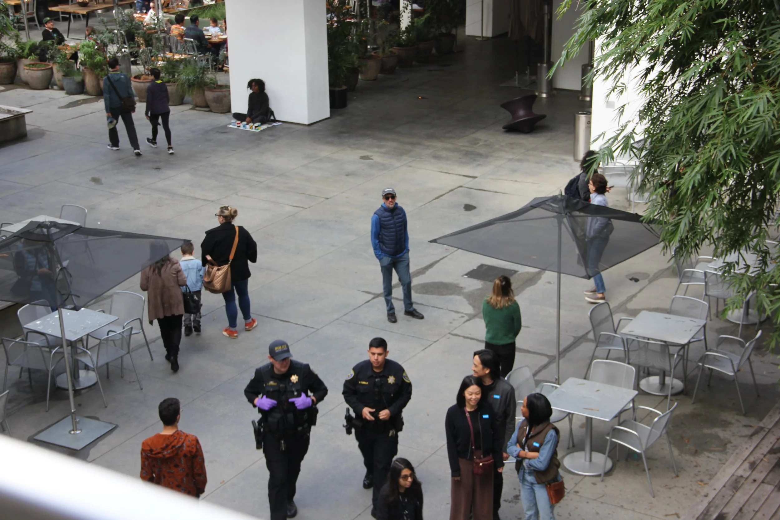 People walking and standing in an outdoor patio area with tables and umbrellas, some police officers are present, and a person sitting on the ground near a white wall.