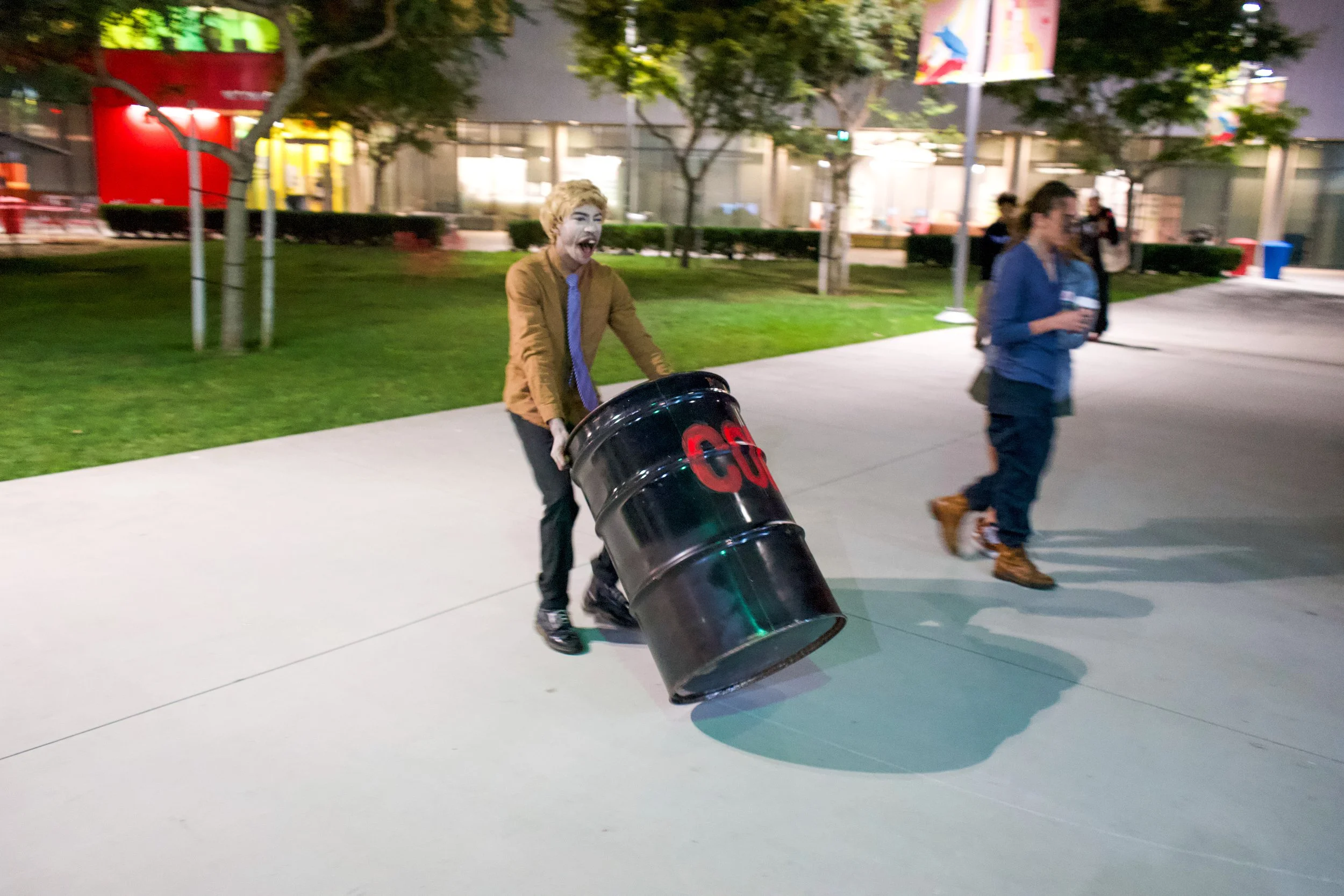A person with blonde hair and a brown jacket pushes a black barrel labeled 'COG' on a sidewalk at night, with two other people walking in the background near trees and modern buildings.