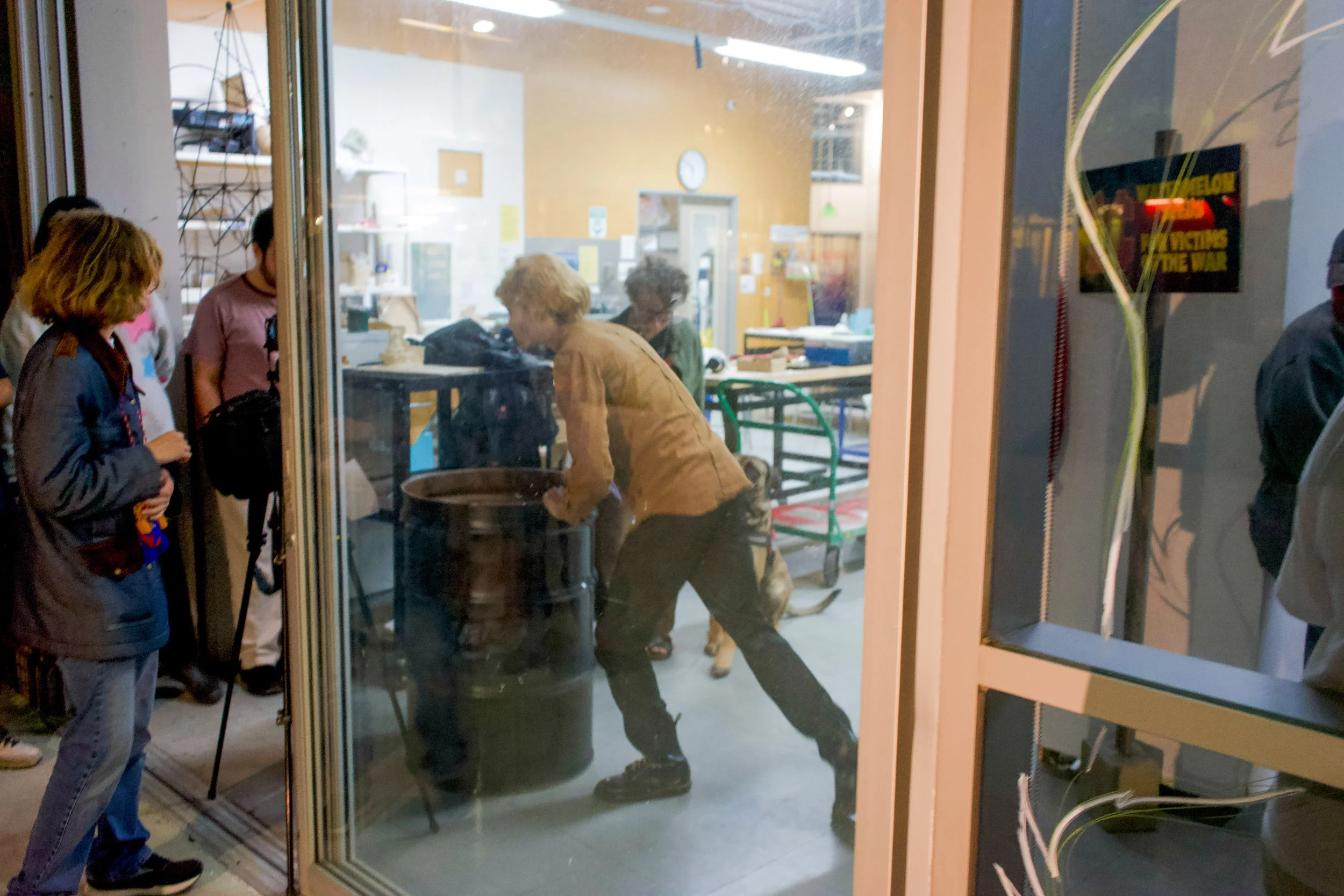 View through a glass door showing several people inside a room with yellow walls. One person appears to be pushing or hitting a trash can while others observe.