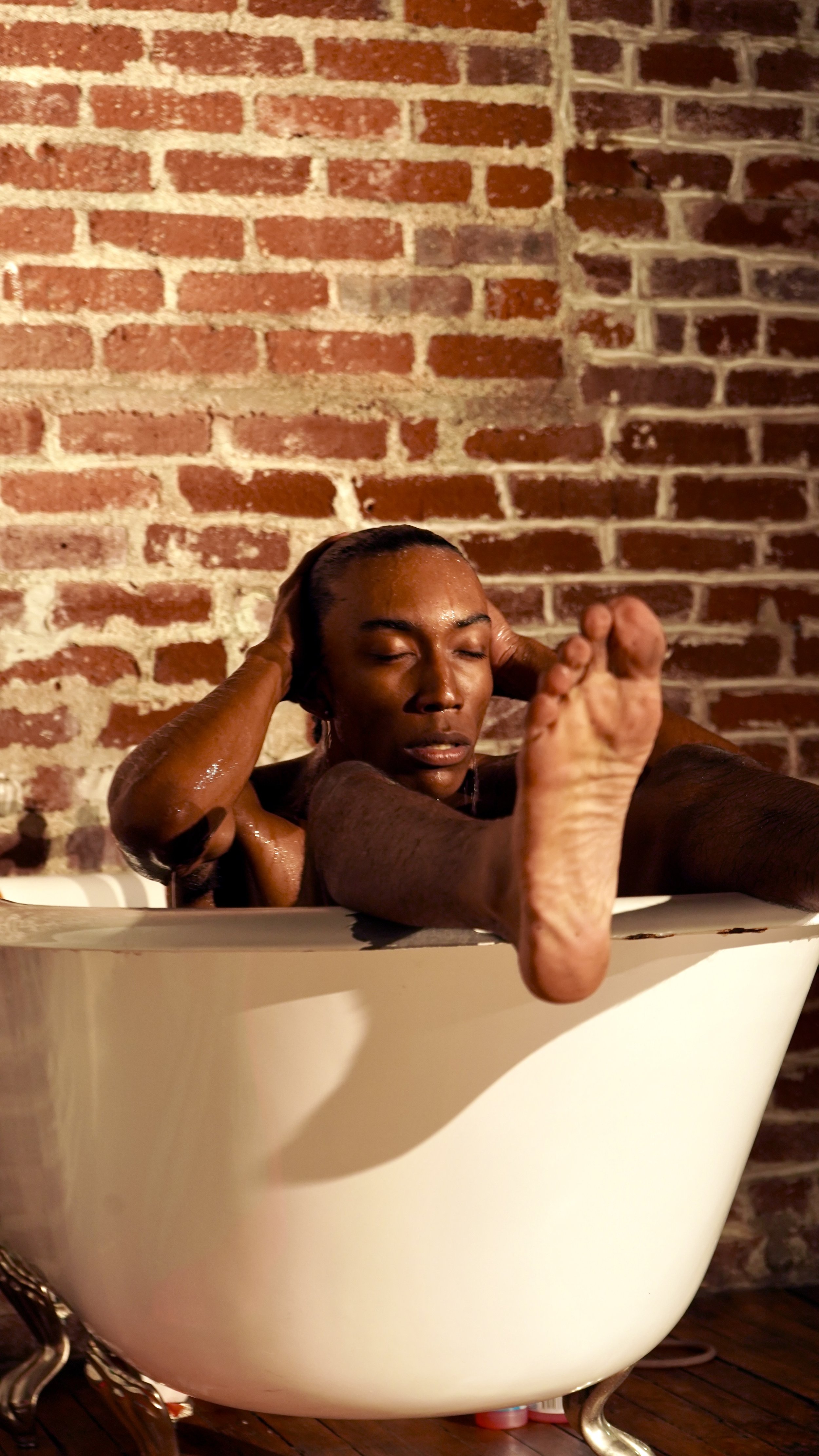 A woman with wet hair relaxing in a white bathtub against a brick wall.