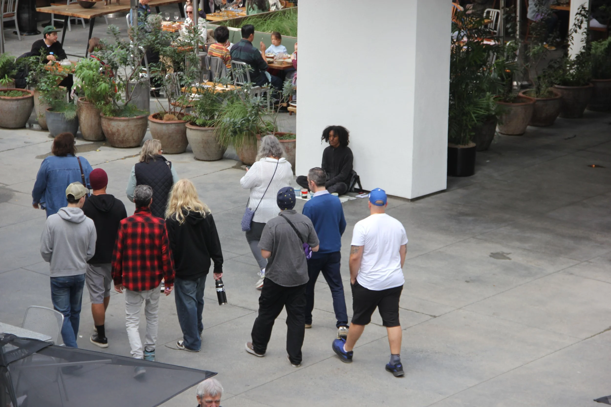 A street performer with curly hair sitting cross-legged against a wall, surrounded by a group of people watching or passing by, with a restaurant area in the background.
