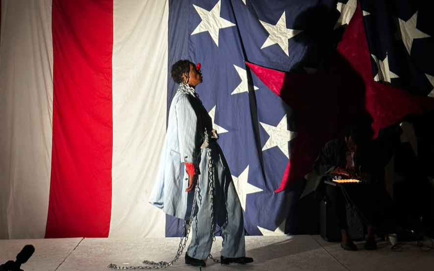 A person standing on stage in front of a large American flag backdrop with red, white, and blue colors and stars; they are wearing chains, a striped suit, and red sunglasses, with a large red star decoration to the right.