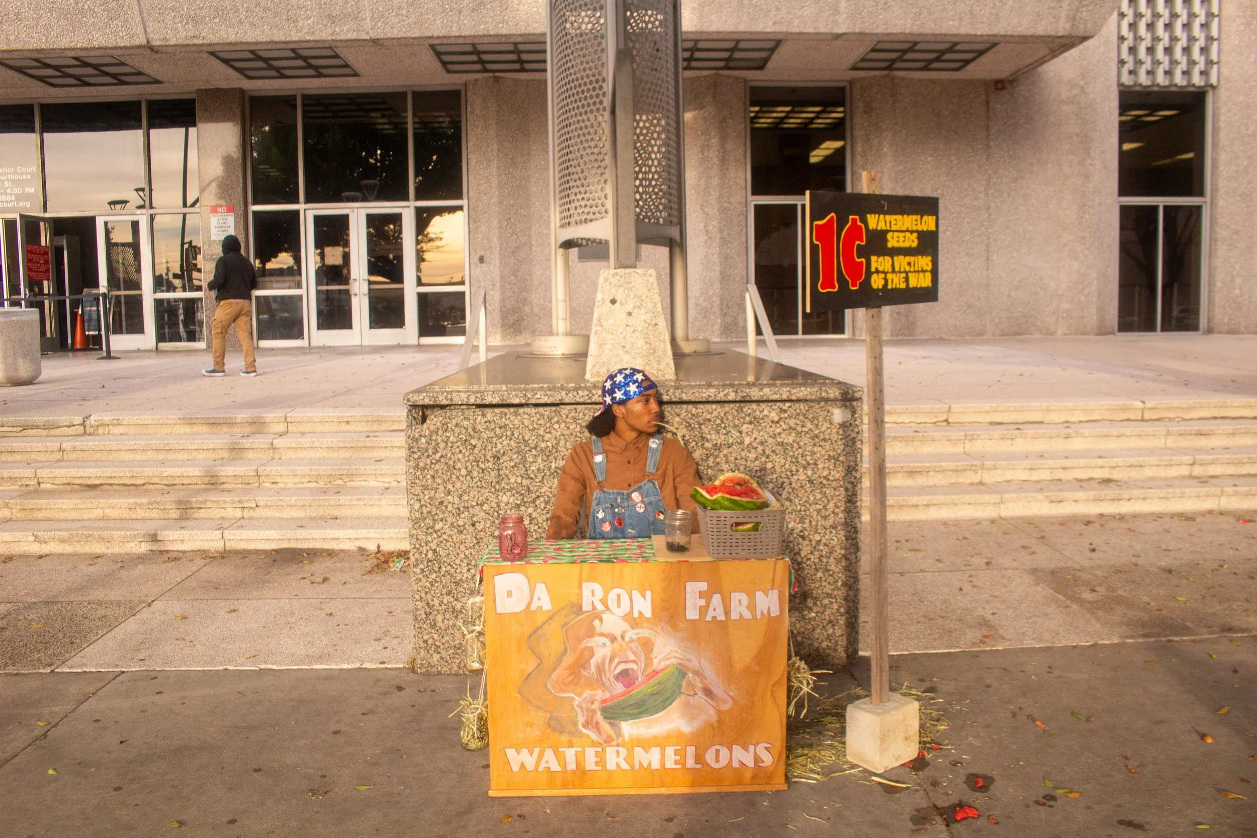 A street vendor selling watermelons at a small stand outside a public building. The stand has a sign that reads 'Daron Farm Watermelons' and a painted image of a woman eating watermelon. The vendor is wearing overalls and a bandana with stars, and si
