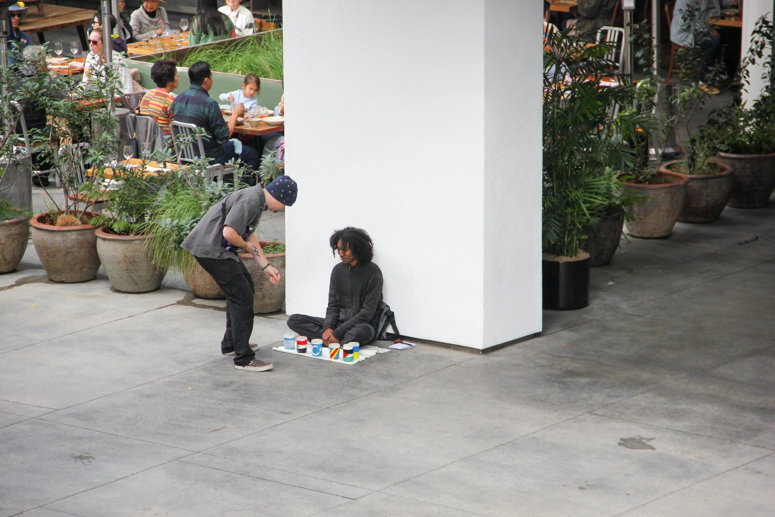 A person sitting against a white wall on the sidewalk with a collection of painted cans and cups in front of them, while a man is crouching nearby, seemingly interacting with them. In the background, there is a busy restaurant with people dining.