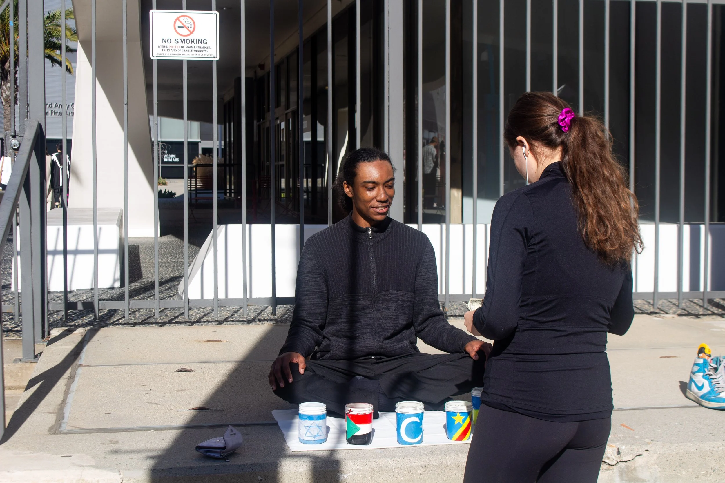 A woman with a purple hair clip and earphones stands in front of a seated man at a sidewalk stand with painted jars. The jars display various symbols and flags, including a Star of David, Palestinian flag, crescent moon, and other colorful designs.