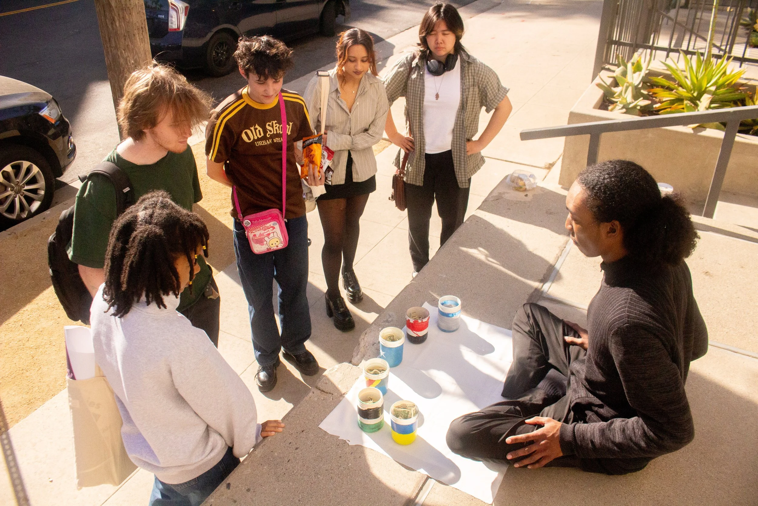 A man is sitting on a ledge with a white sheet in front of him, displaying multiple folded banknotes, while a group of young people stands around, looking at the money.