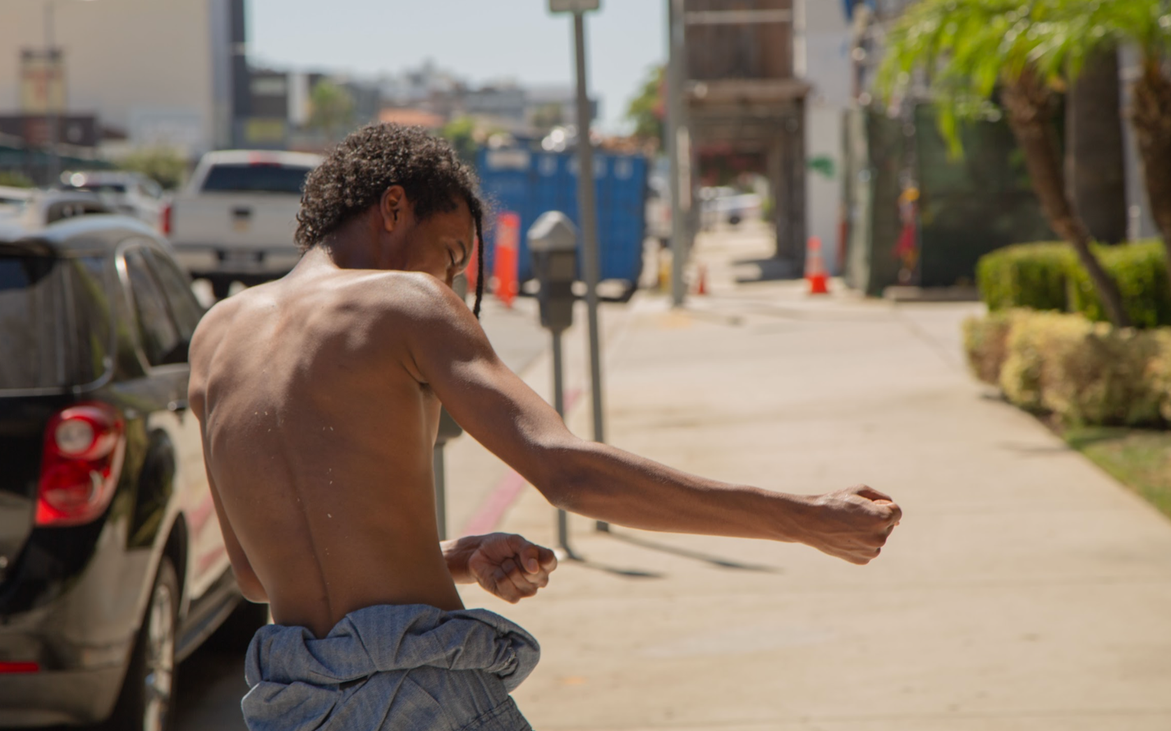 A shirtless man with curly hair, seen from the back, is outdoors on a sunny day, wearing gray pants around his waist, and appears to be flexing or engaging in a stretching pose on a sidewalk.