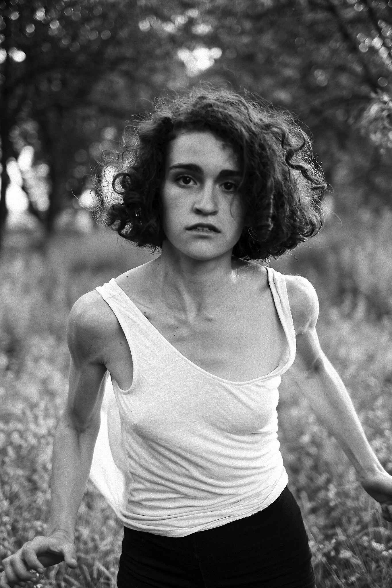 Black and white photo of a woman with short curly hair, wearing a tank top, outdoors with trees in the background.