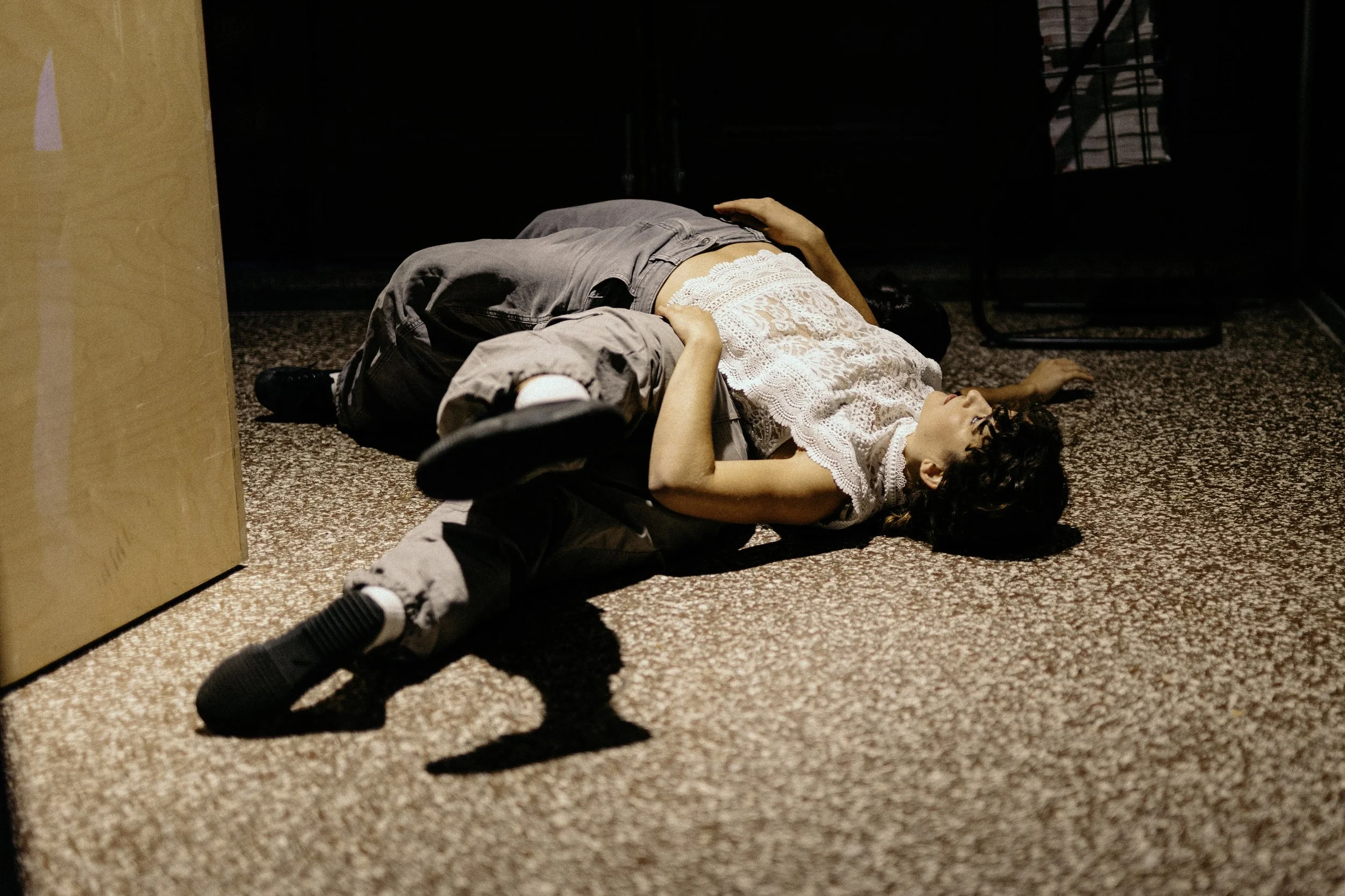 Two people lying on the floor, one with a white lace top, the other in gray pants and a gray jacket, in an indoor space with a carpeted floor.