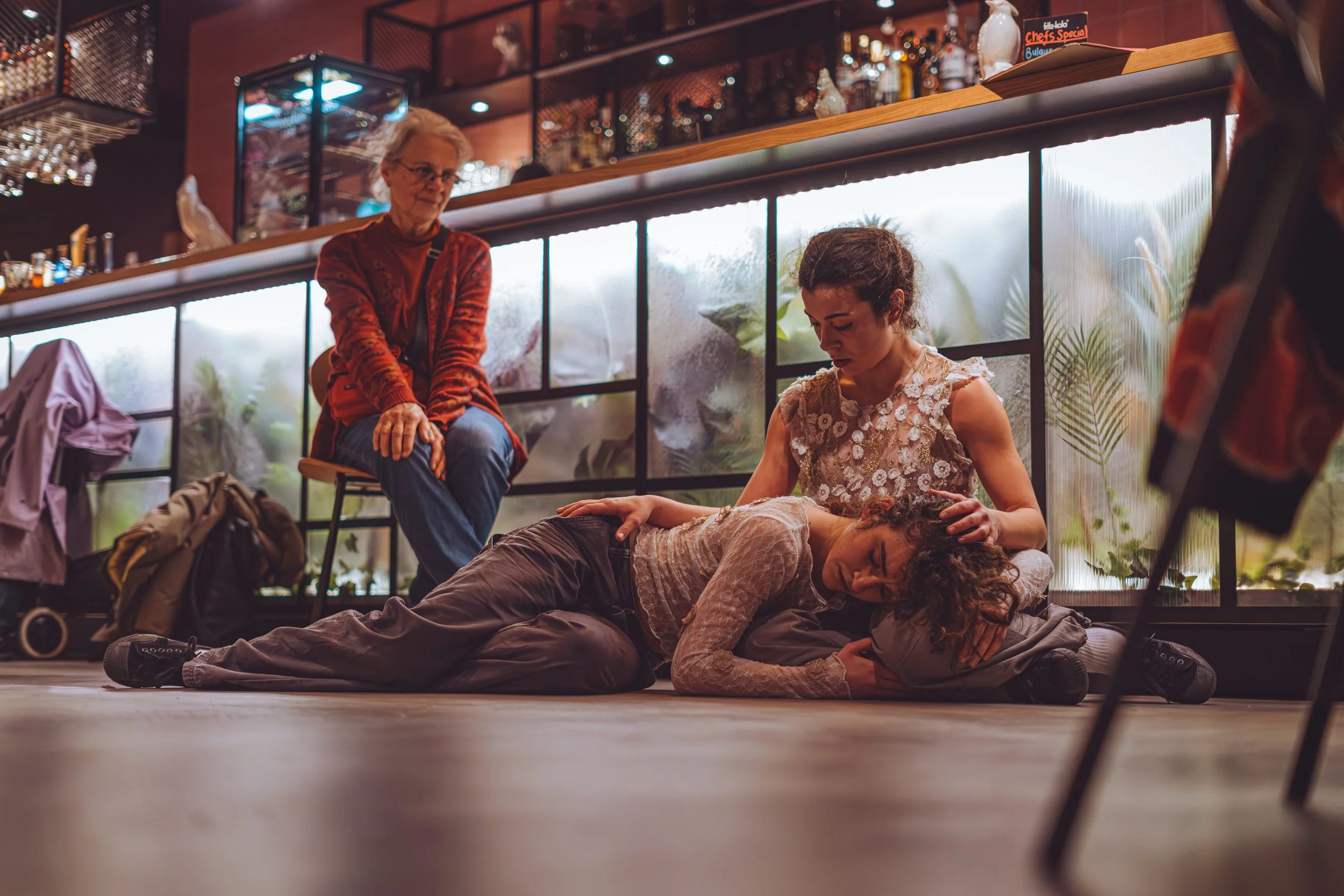 A woman lies unconscious on the floor with a woman kneeling beside her, gently holding her head and shoulder. A third woman sits nearby on a barstool, observing the situation with concern. The setting appears to be a restaurant or cafe, with large fr