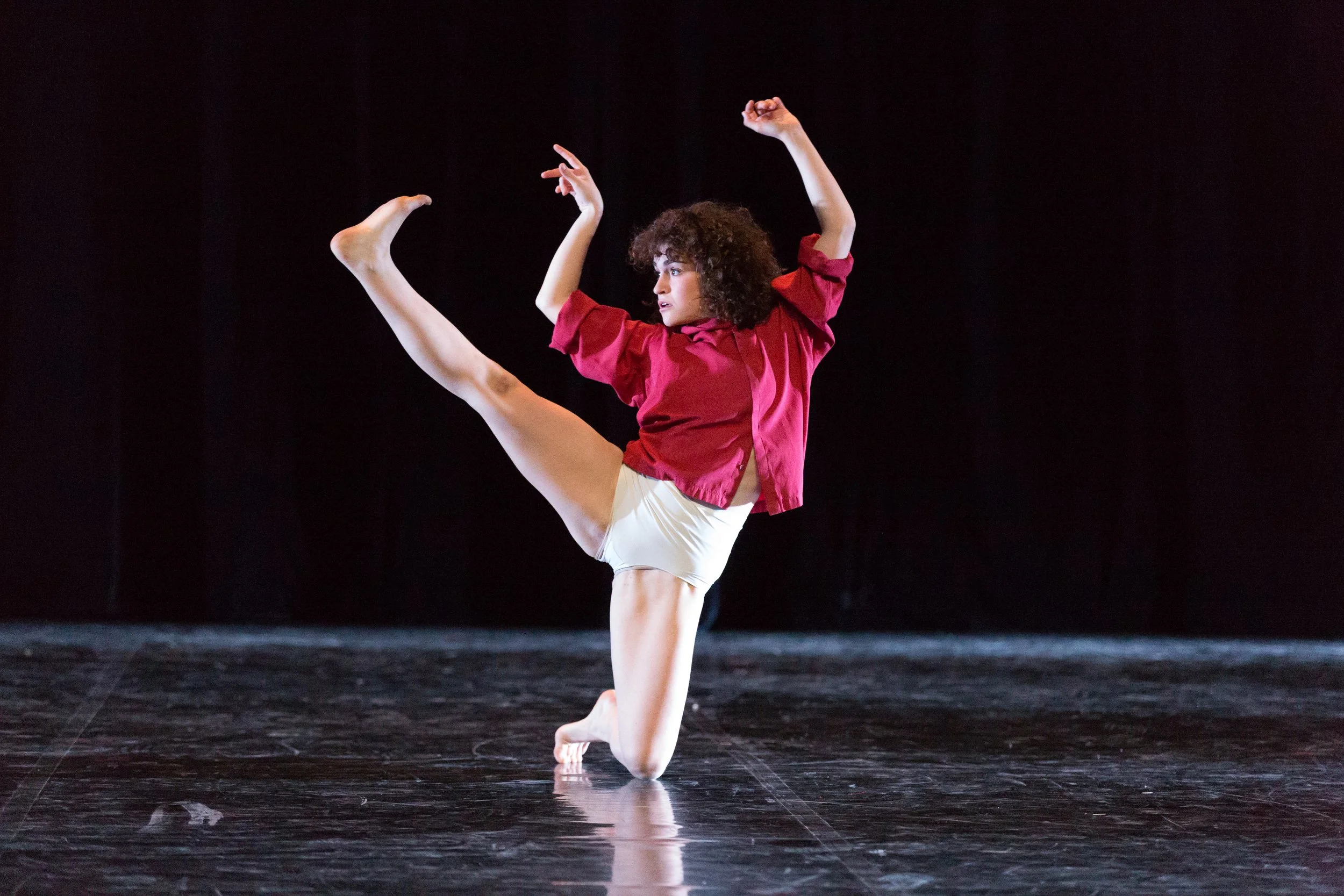 A woman performing a dance move on stage, wearing a red shirt and white shorts, with a black background.