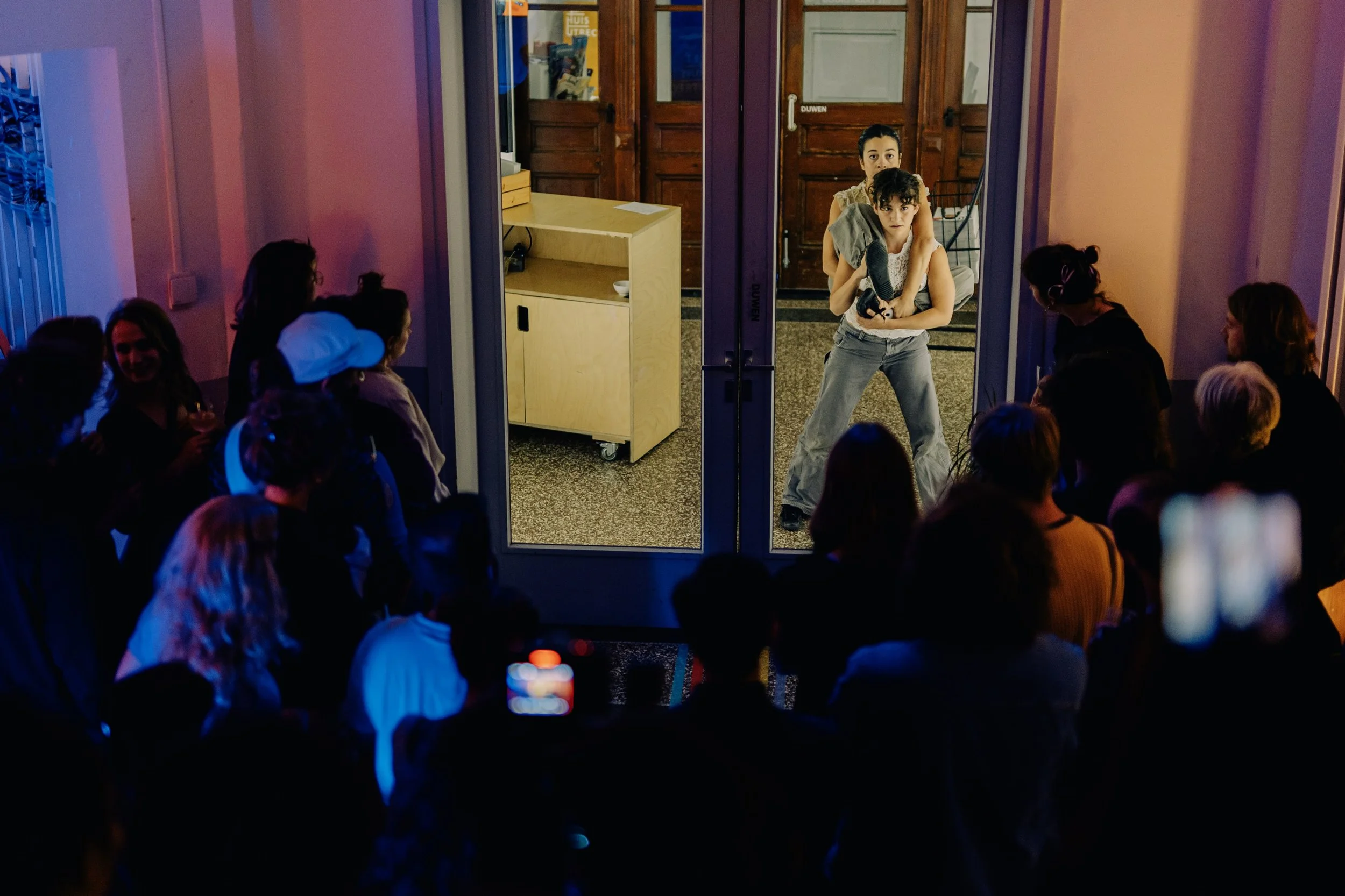 Two young women are performing for an audience of mostly women in an indoor venue. The performers are standing at the entrance of a room, with one holding a microphone and the other holding a microphone stand. The audience is seated and watching atte