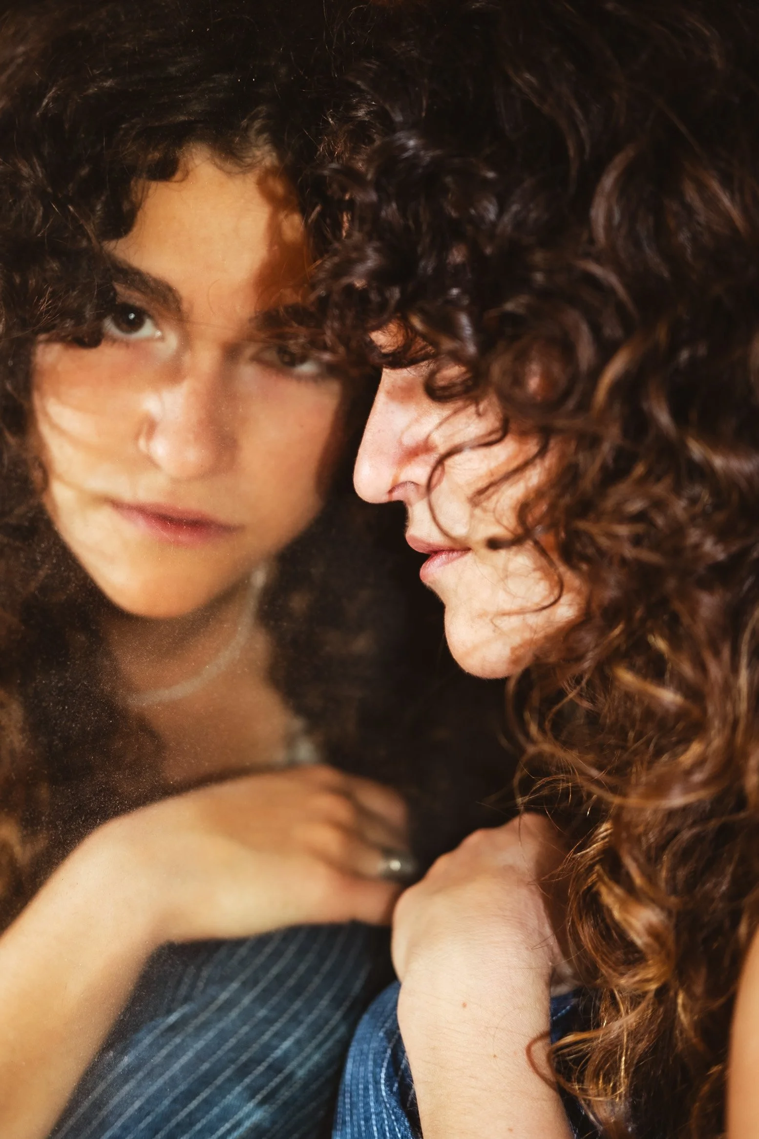 A woman with curly hair looks at her reflection in a mirror, with her face close to the glass and her hand resting on her chest.