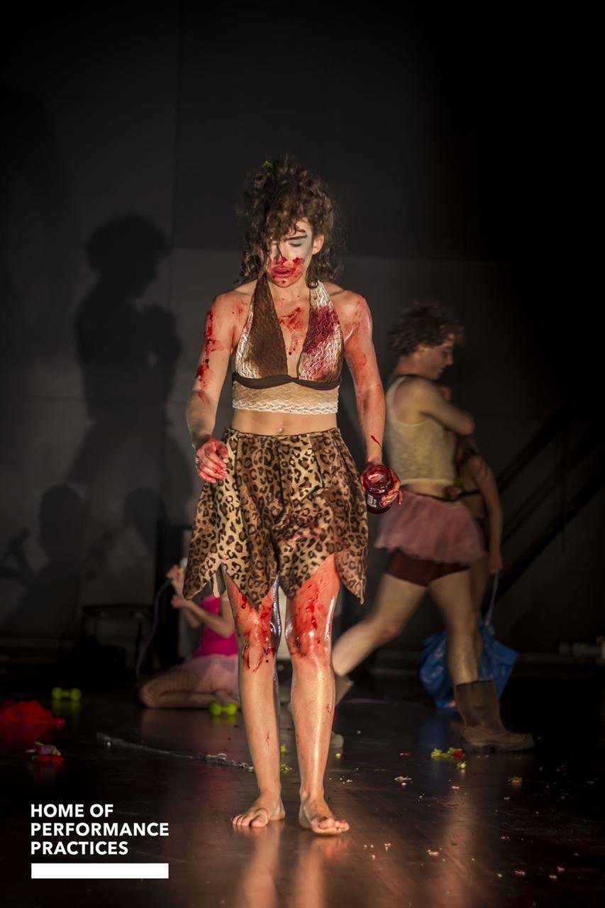 Performer covered in fake blood, wearing leopard print skirt and a bra top, on stage with other performers, backstage or rehearsal setting.