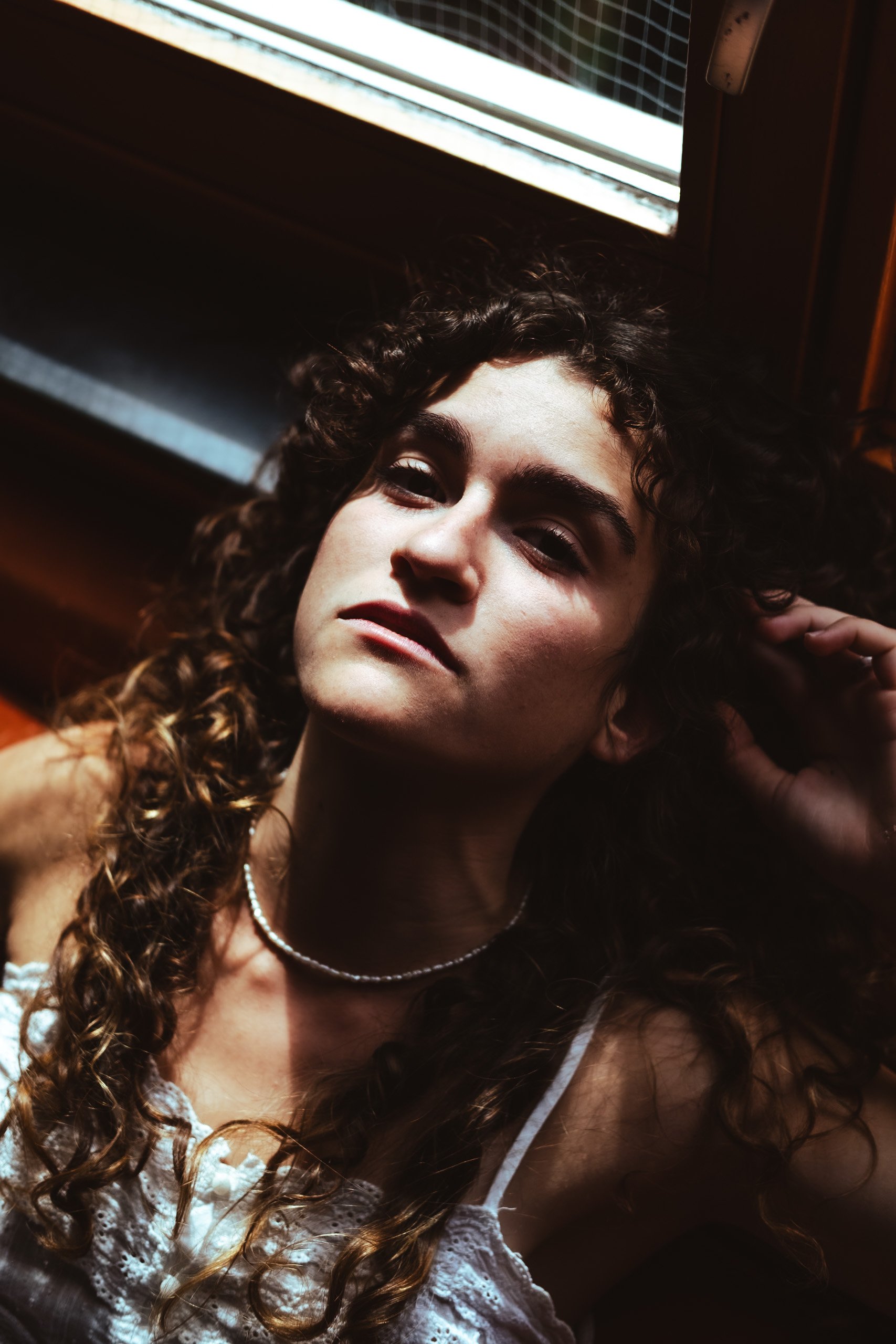 Young woman with curly hair lying near a window, looking at the camera with a serious expression.