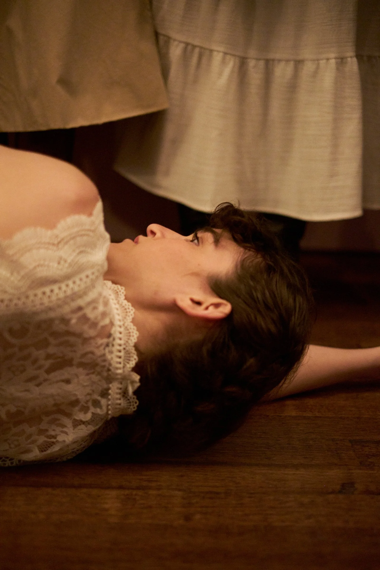 A young woman lies on a wooden floor, looking upwards, with her arm stretched above her head. She has short, wavy dark hair and is wearing a white lace top. There are curtains in the background.