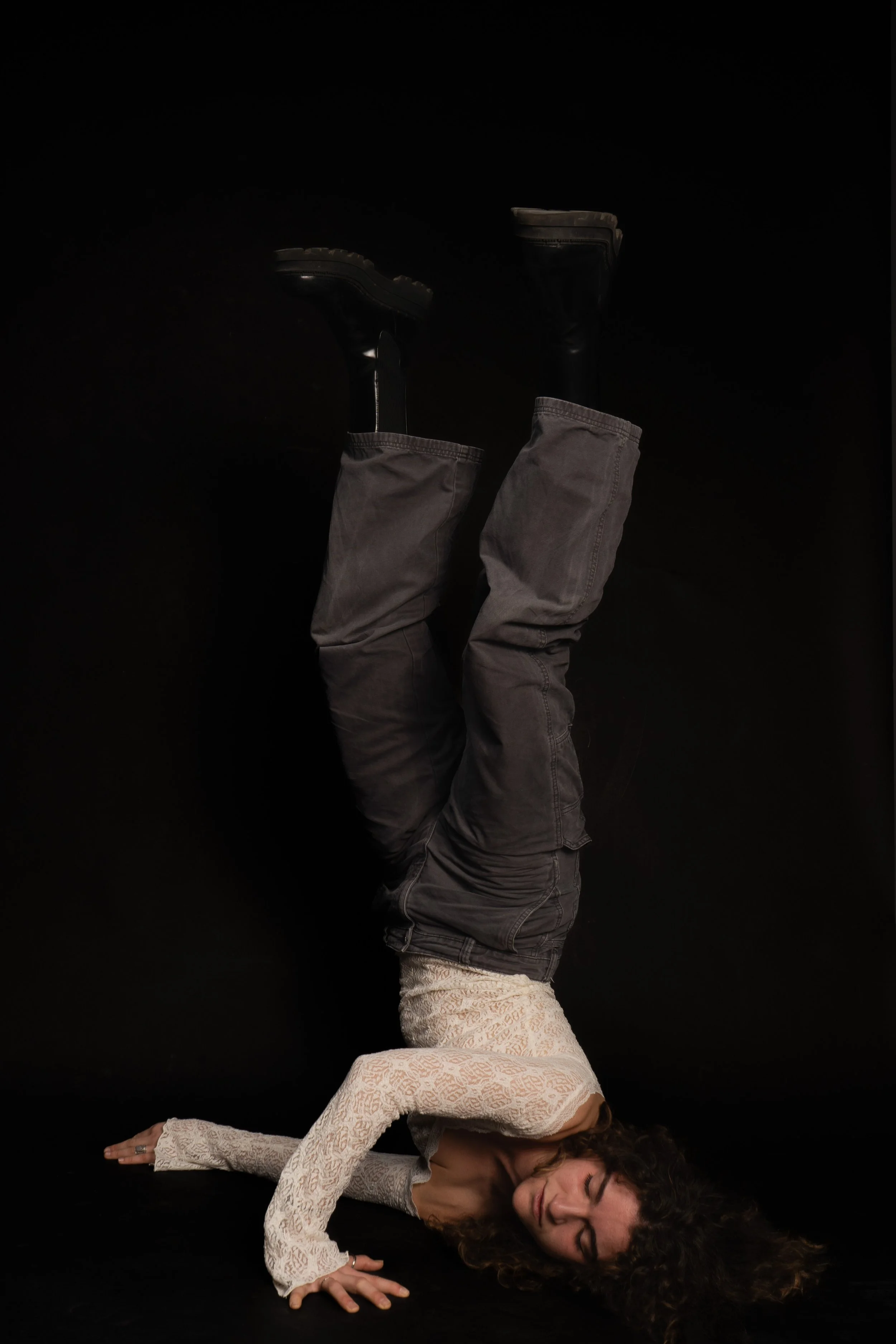 Woman lying on the floor in a handstand pose against a black background, wearing a white lace top and dark pants.