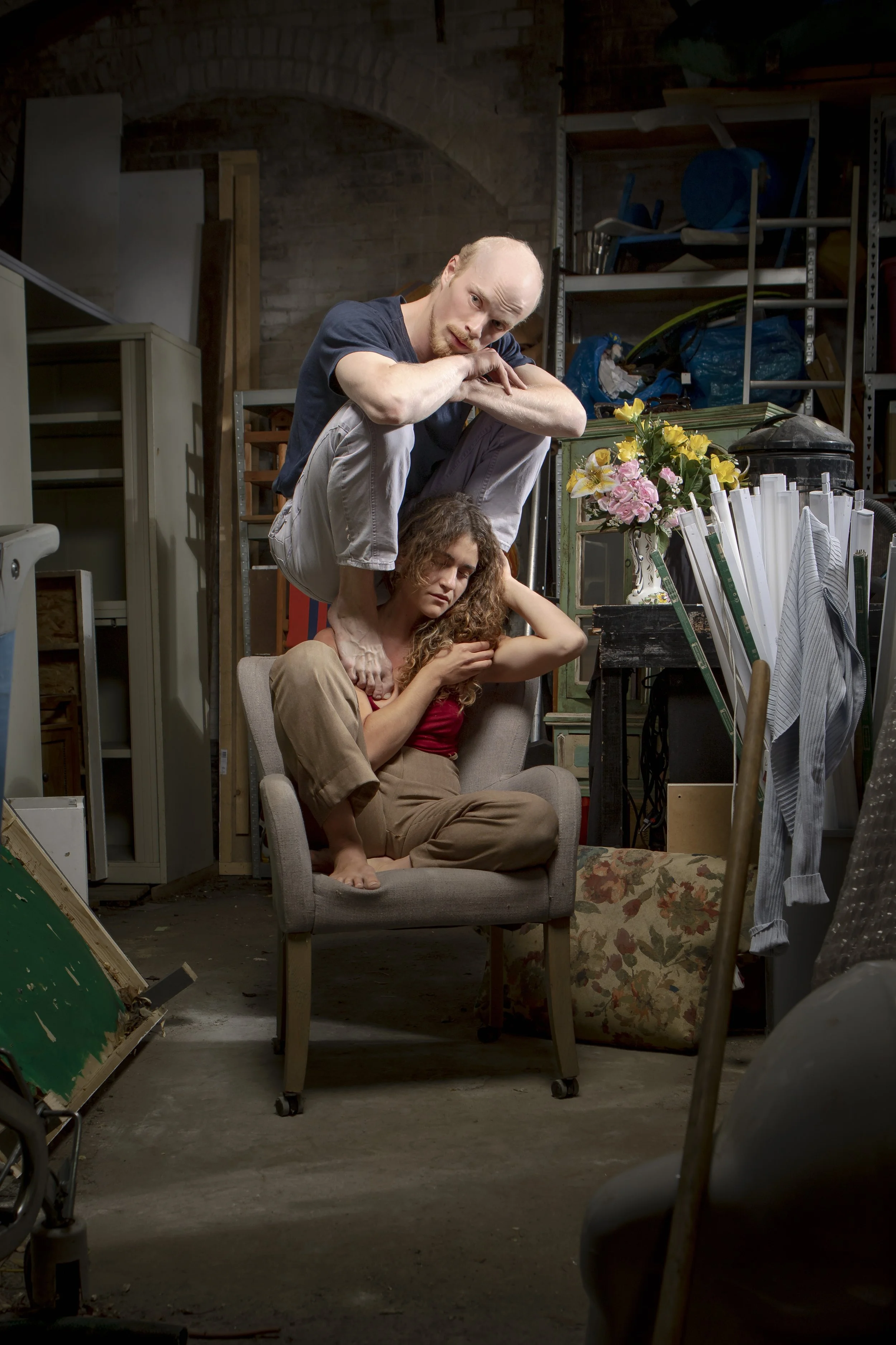 A man and a woman are posed in a cluttered garage, with the man sitting on a chair with one knee up and leaning over the woman, who is sitting on the chair with her arms crossed, looking distressed.