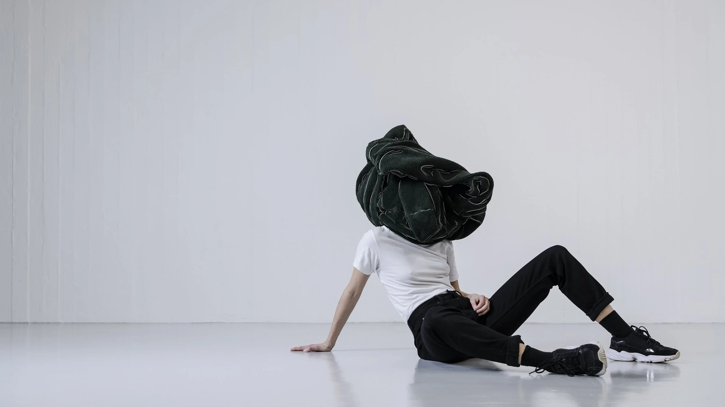 Person sitting on the floor in a white t-shirt and black pants with a large, black, padded bag covering their head, in a minimalistic white room.