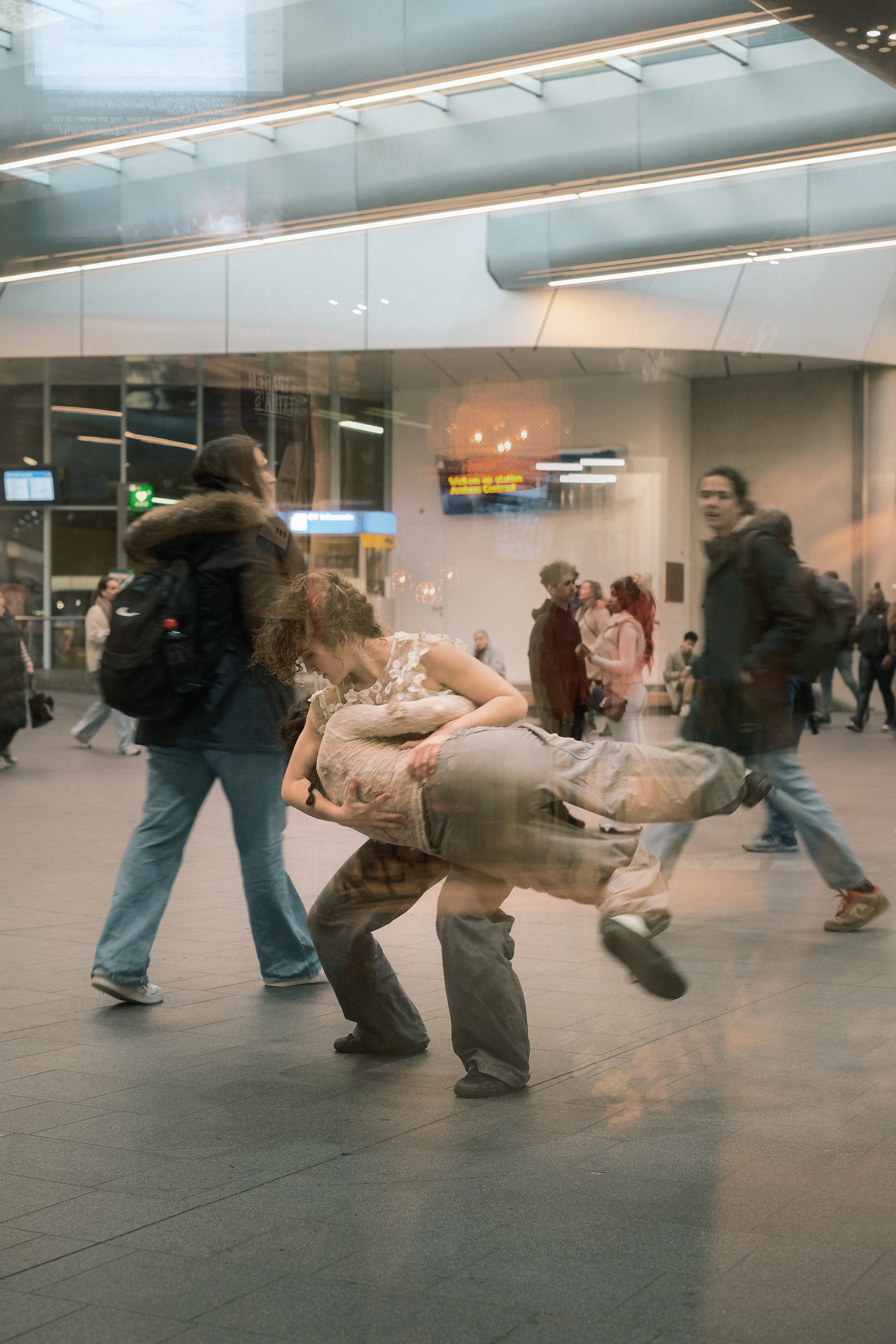 A woman with curly hair wearing a sleeveless lace top is carrying another person, possibly a child, through a busy public space with many pedestrians. The scene appears to be viewed through a glass window or reflection.