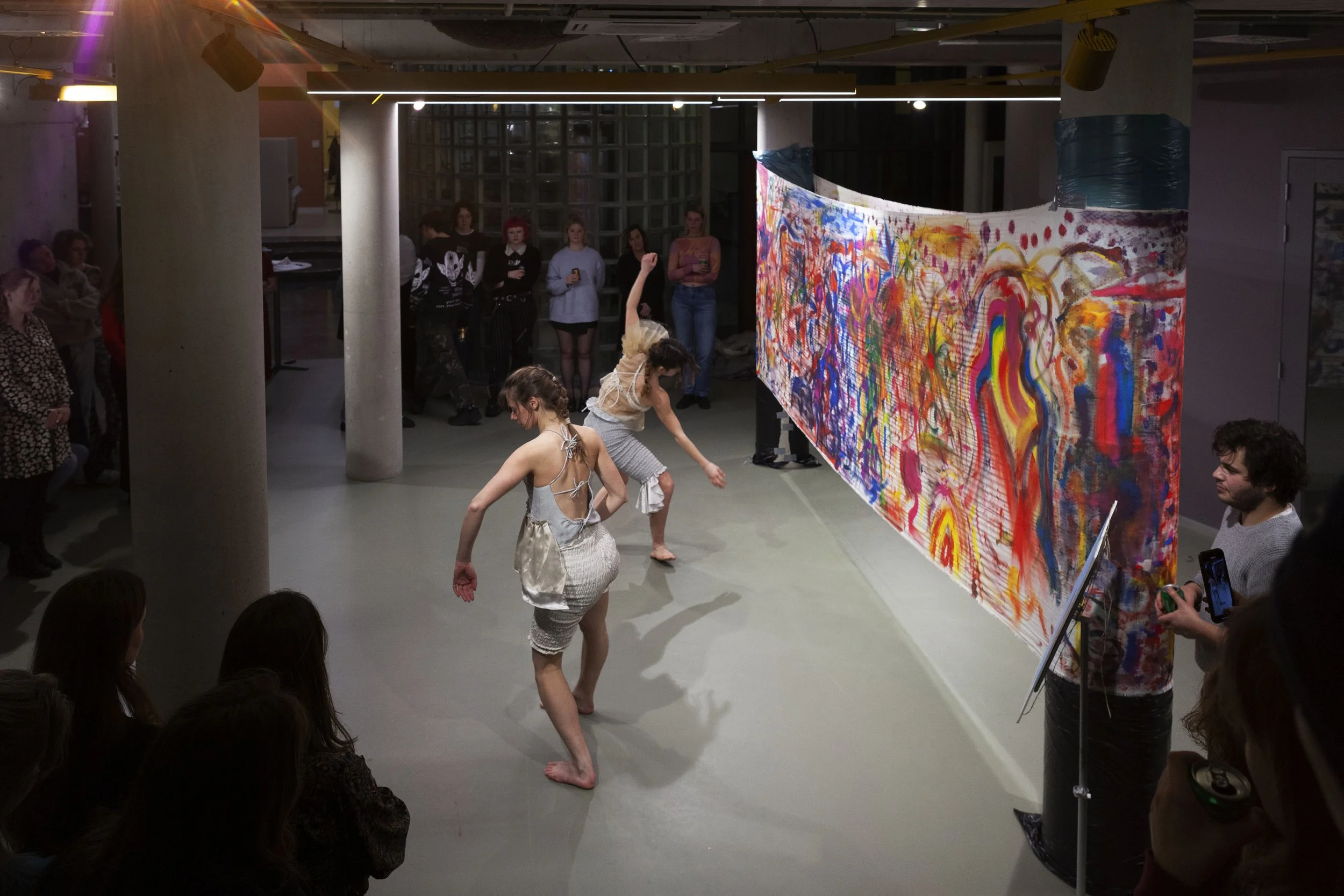Two dancers perform in front of a colorful abstract painting in an art gallery with an audience observing.