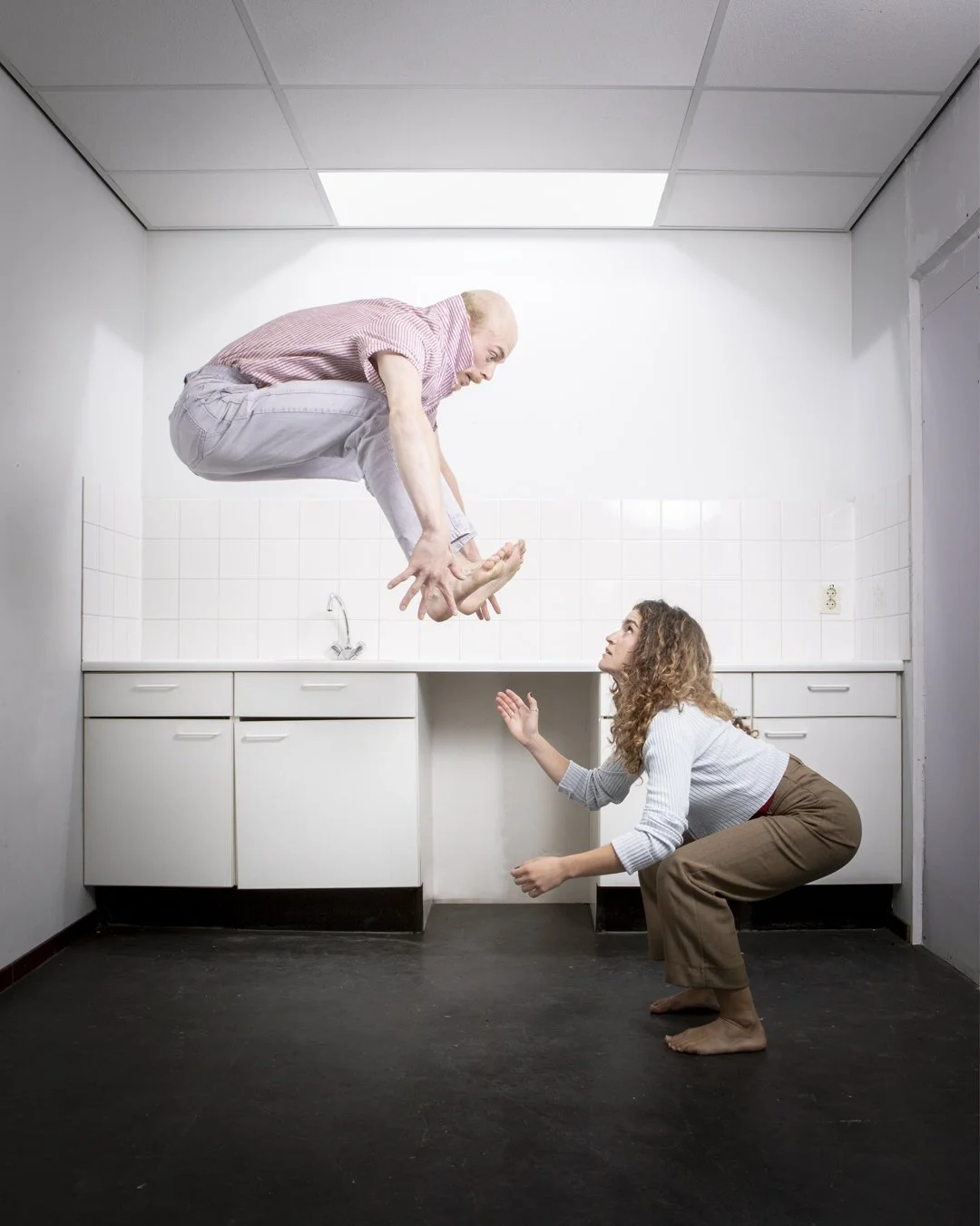 A man and woman in a kitchen with white cabinets and tiled backsplash, the man is mid-air in a crouched position reaching toward the woman, who is crouched on the floor looking up at him with a surprised expression.