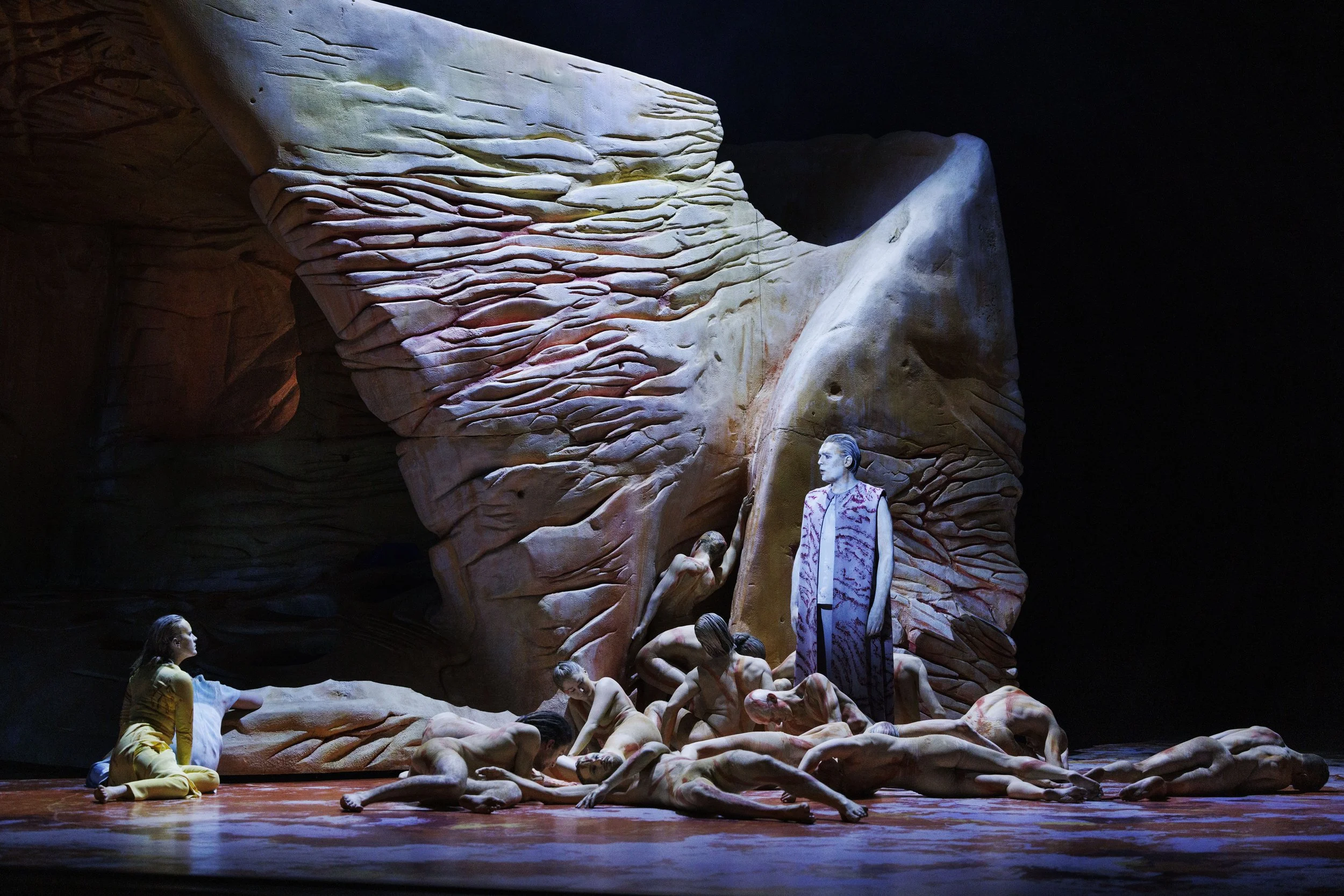 A theatrical stage scene with a large, textured rock wall backdrop. Several actors with painted bodies are on the floor, some kneeling and crawling, while one actor in colorful attire stands upright. One woman in yellow is seated on the floor looking