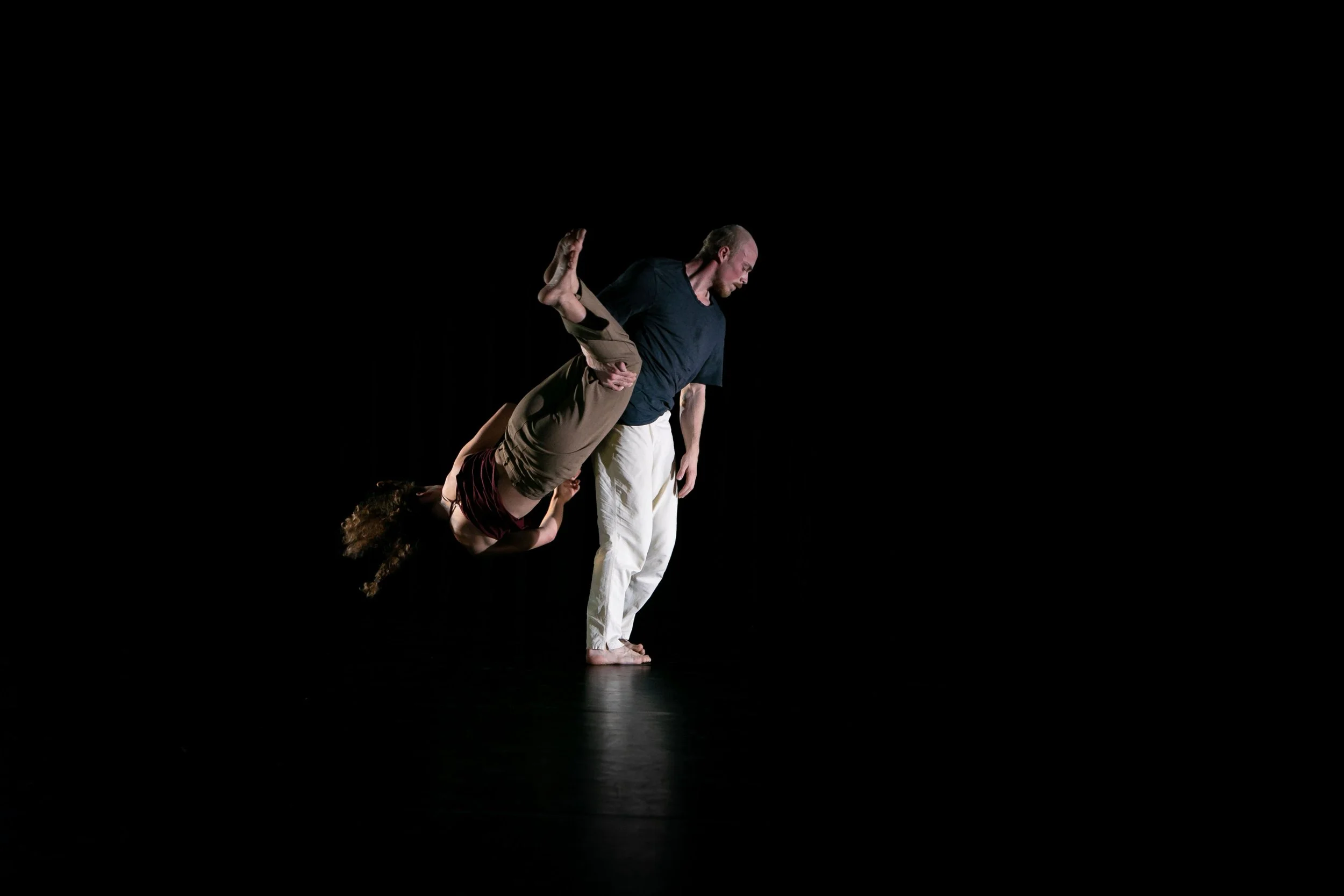 A man in a black shirt and white pants holds a woman in a maroon top and beige pants upside down in a dance pose on a dark stage.