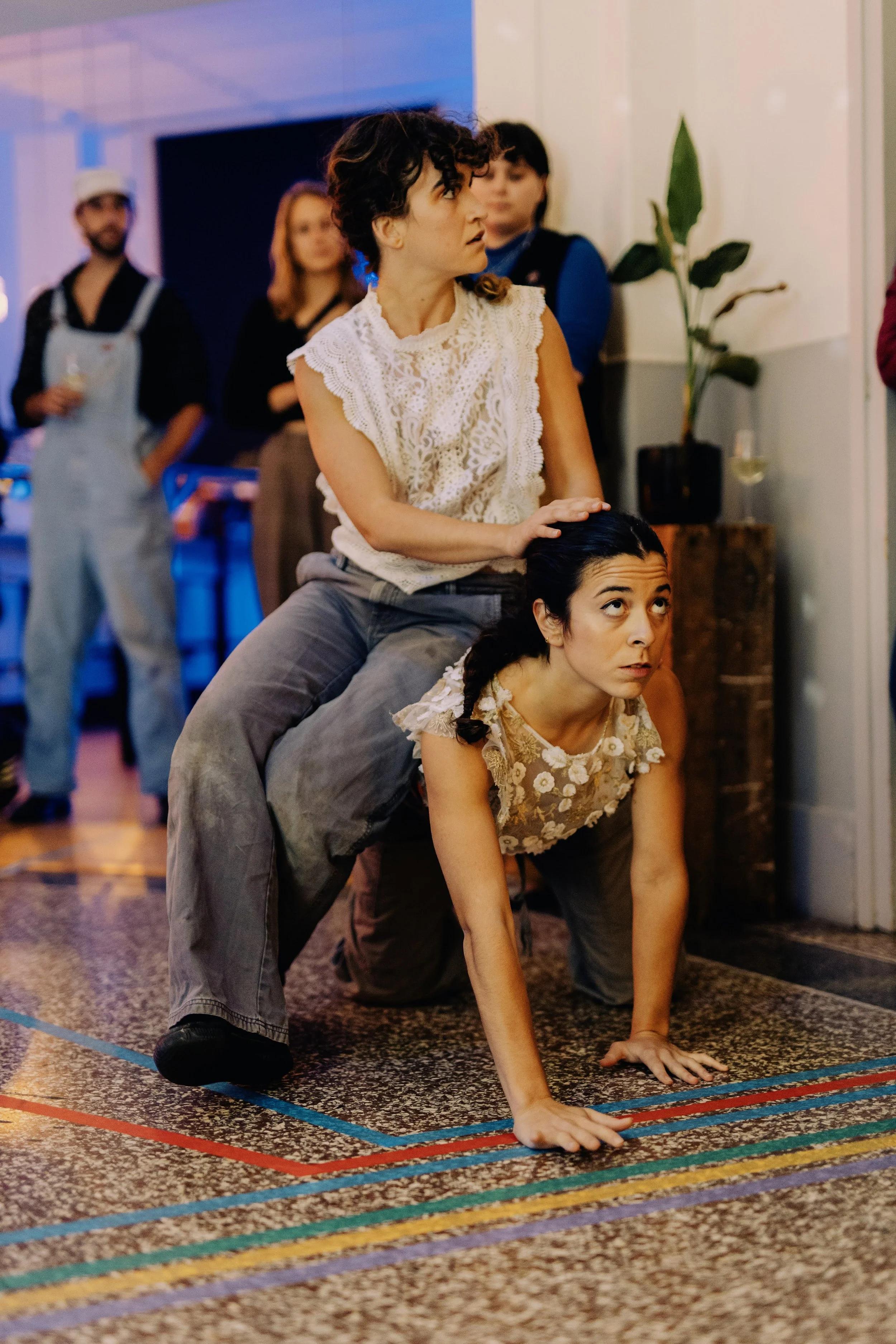 Two women in a social setting, one sitting on the other's back while on her hands and knees, with several people in the background and colorful lighting.