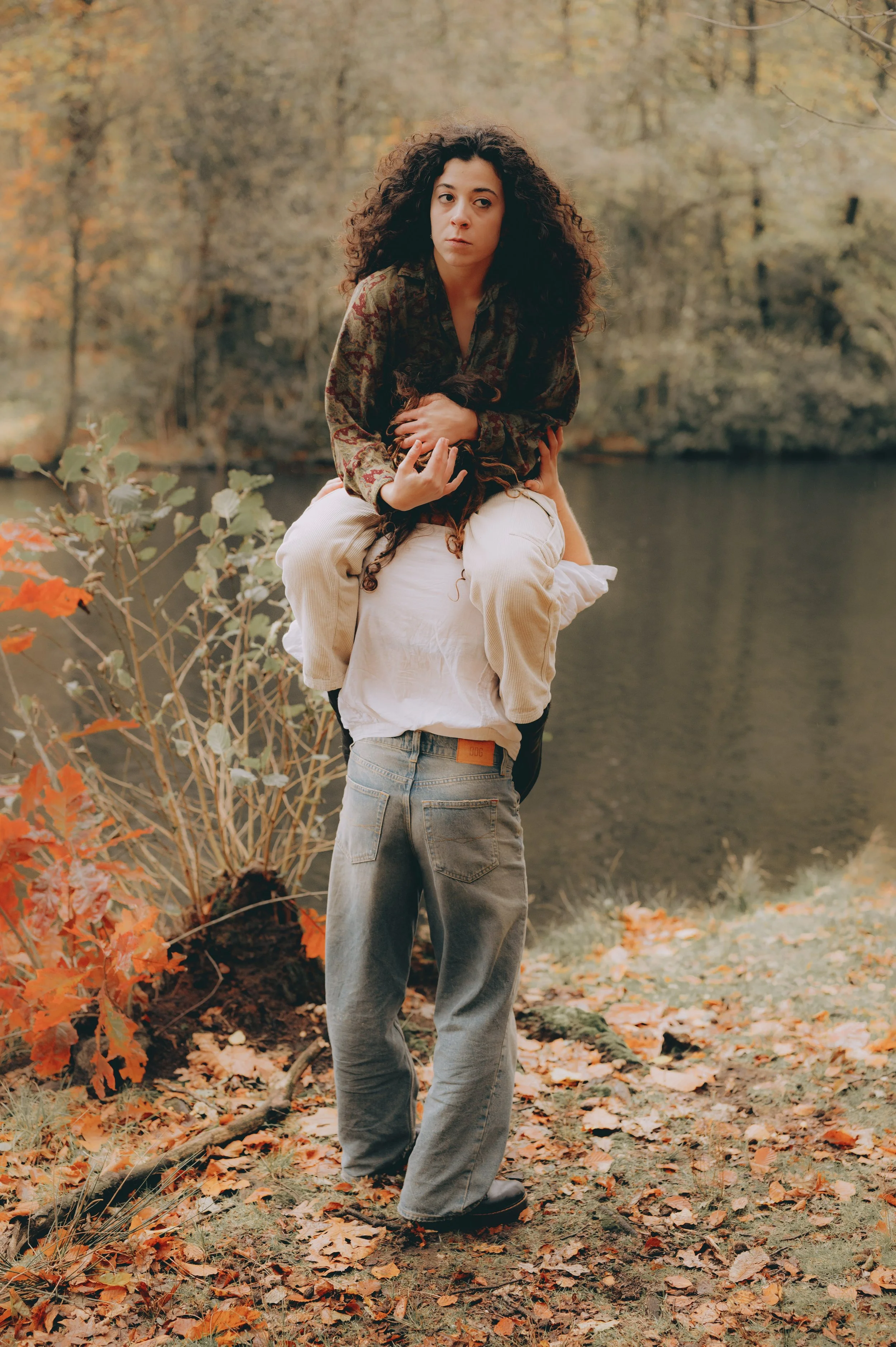 A woman with long curly hair sitting on the shoulders of a man in a park during fall, with autumn leaves on the ground and water in the background.