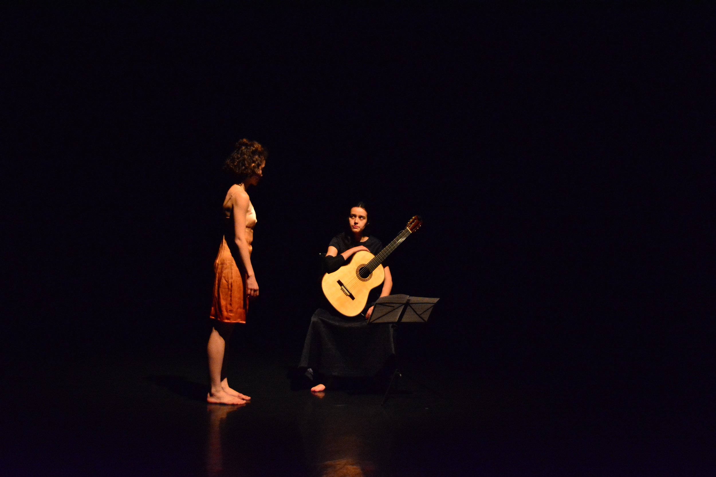 Two women on a dark stage, one standing and the other sitting with a guitar and music stand.