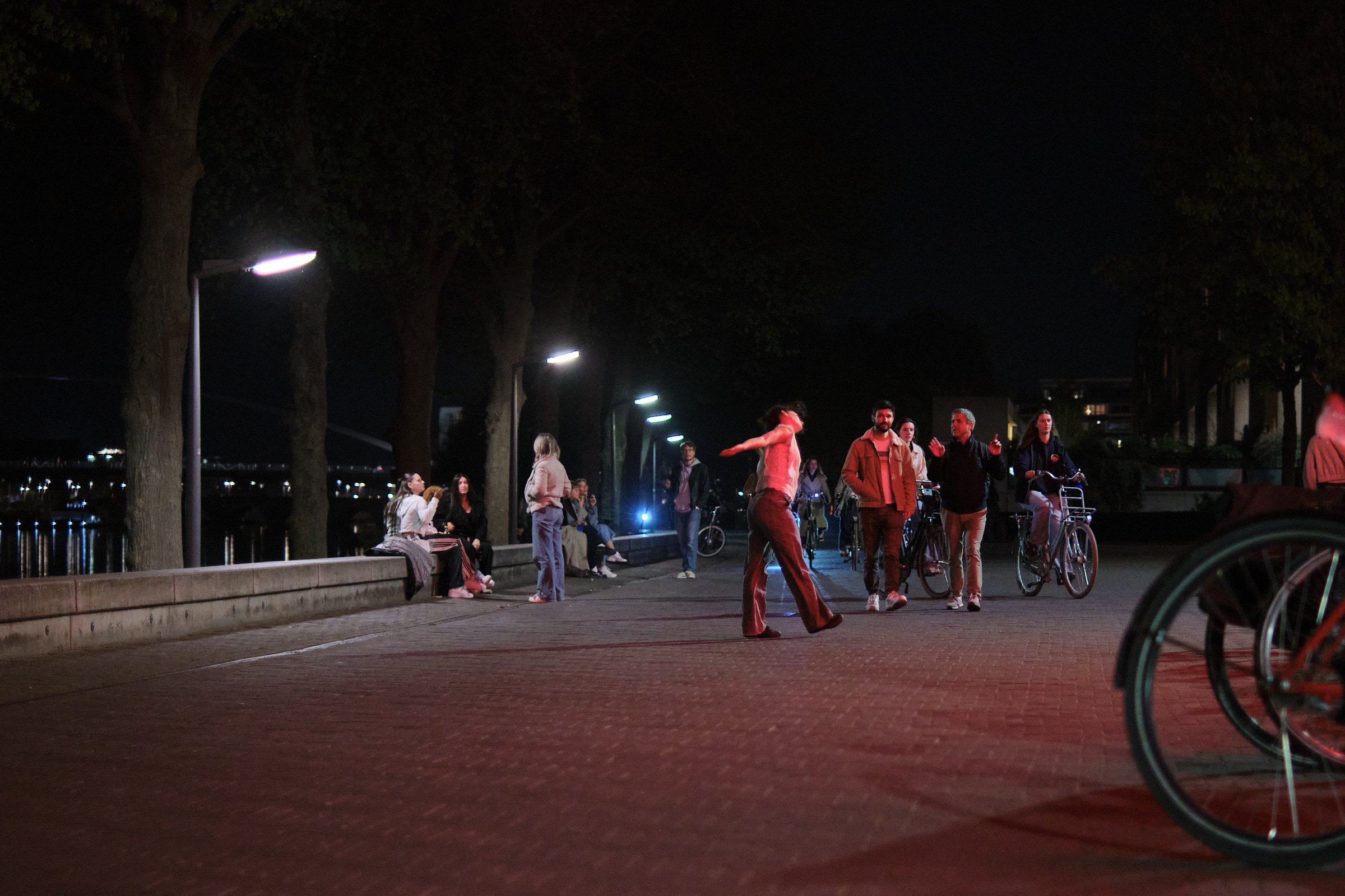 People gathering and socializing in a park at night, some sitting on a bench, others standing or walking, with bicycles nearby and illuminated streetlights along a pathway.