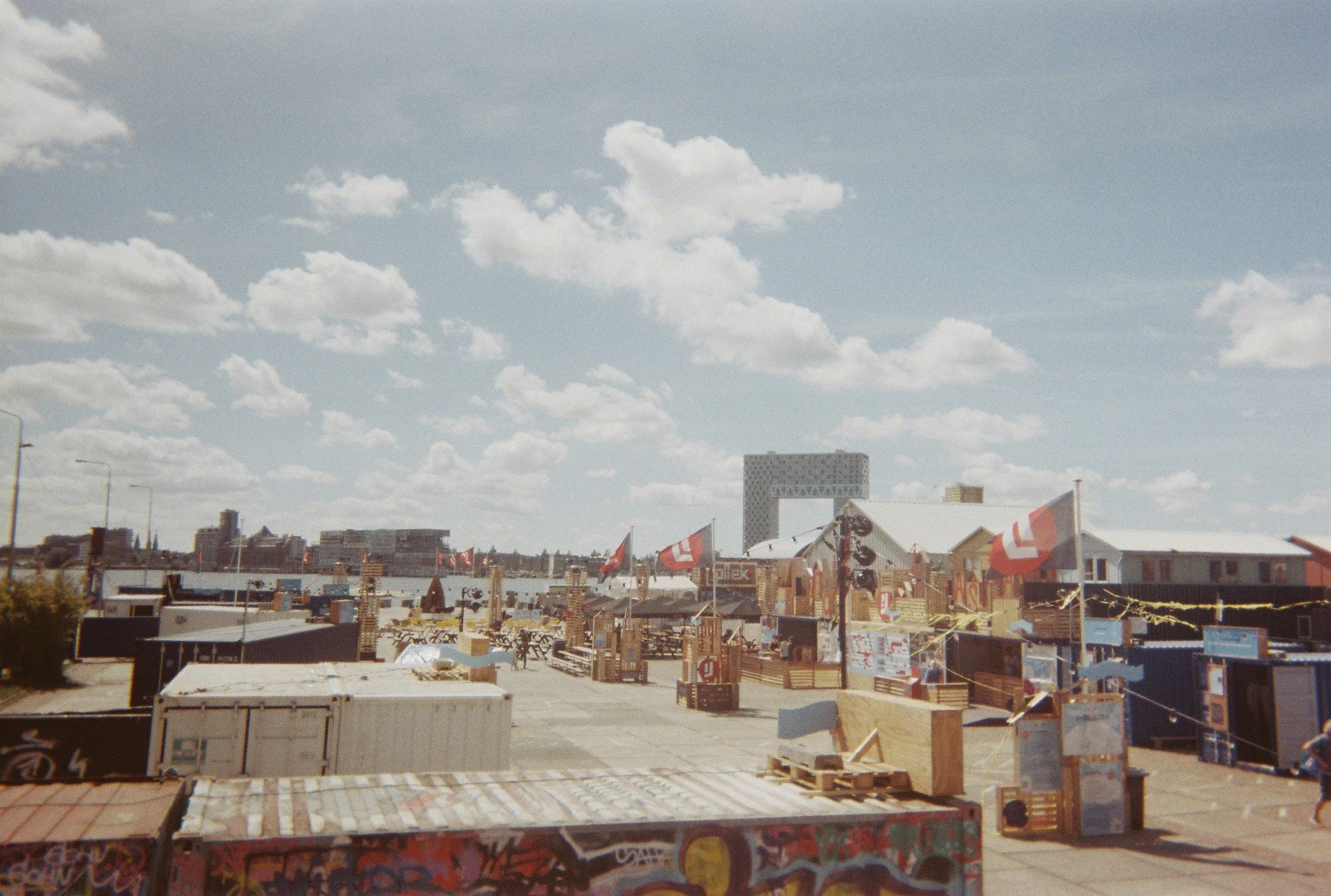 Outdoor cityscape featuring temporary structures, flags, and graffiti, with a cloudy sky and distant buildings in the background.