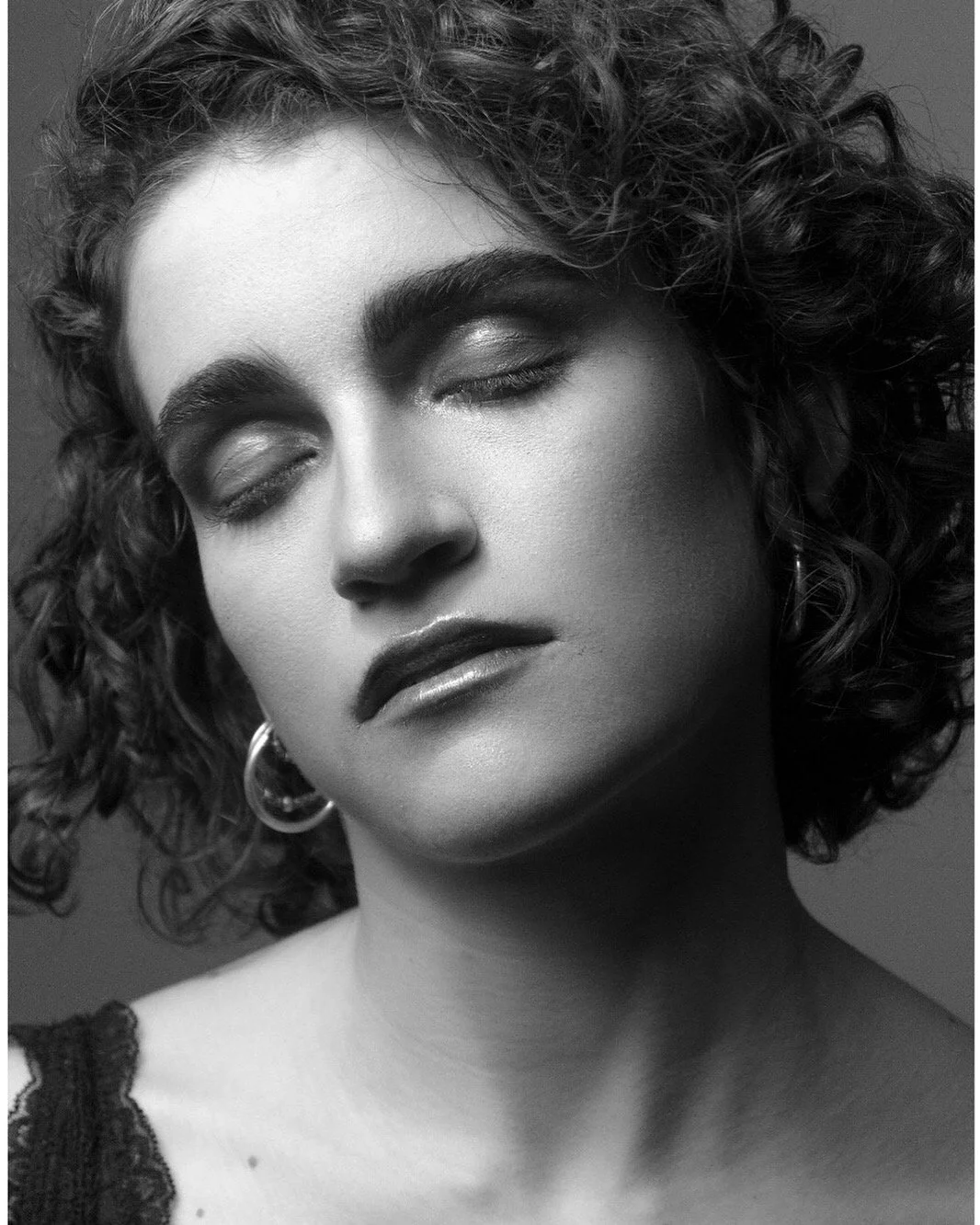 A black and white close-up portrait of a woman with curly hair, closed eyes, and a serene expression. She is wearing hoop earrings and a lace top visible on her shoulder.