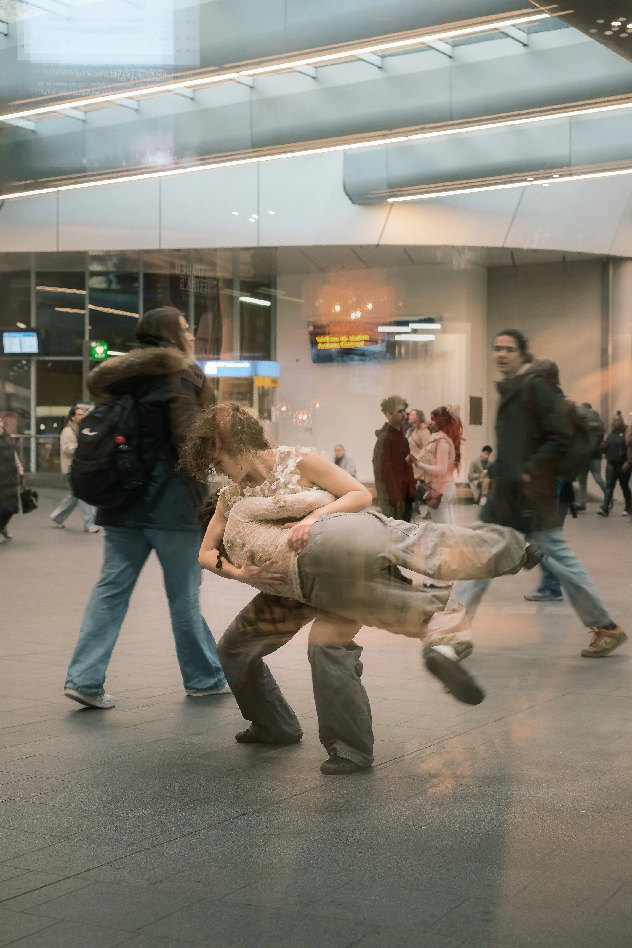 A person is lifting another person in an train station, with other travelers walking by in the background.