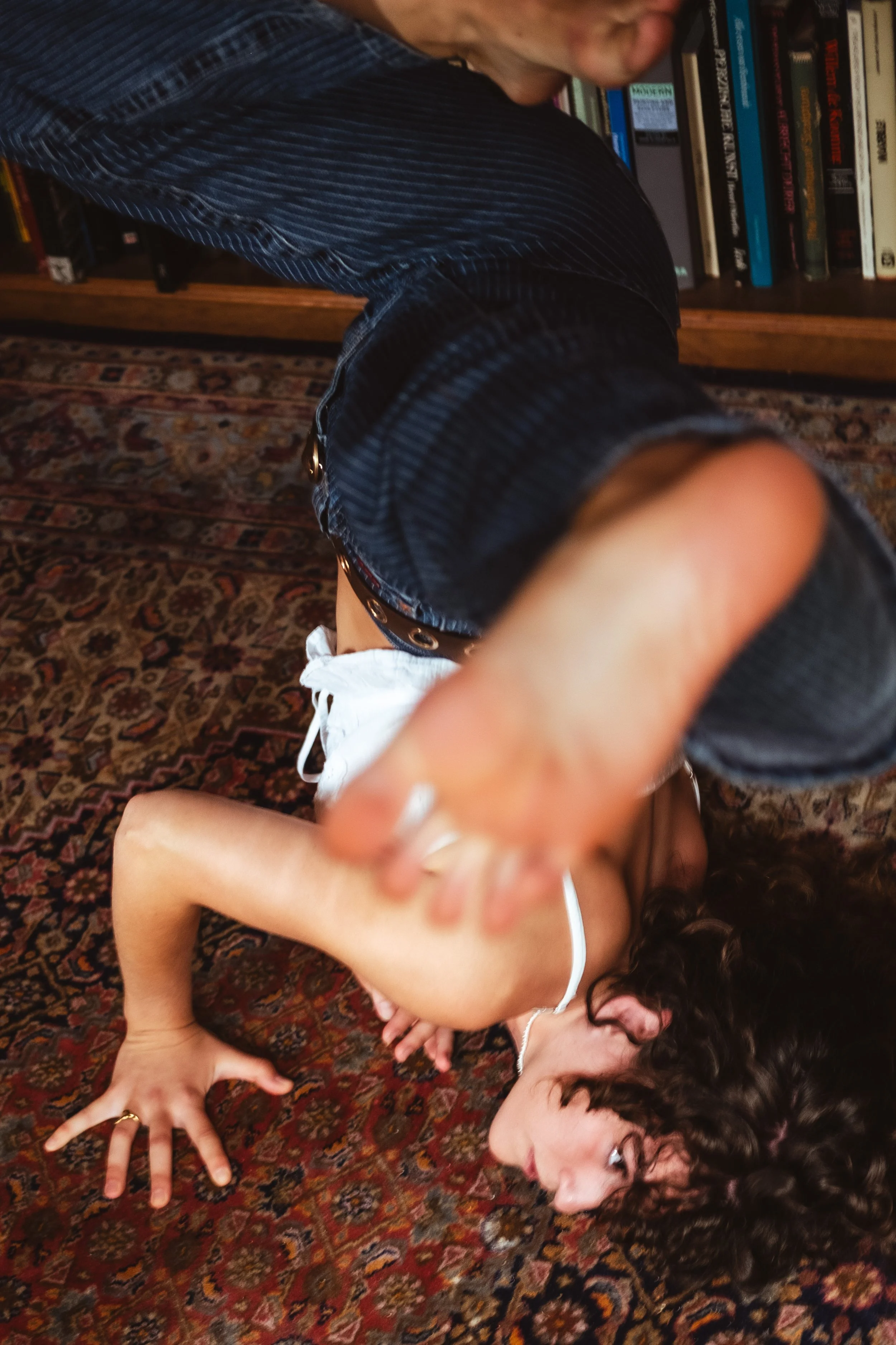 A woman with curly hair lying on a patterned rug on the floor, reaching towards the camera with a somewhat playful or surprised expression, while books are visible on a shelf in the background.