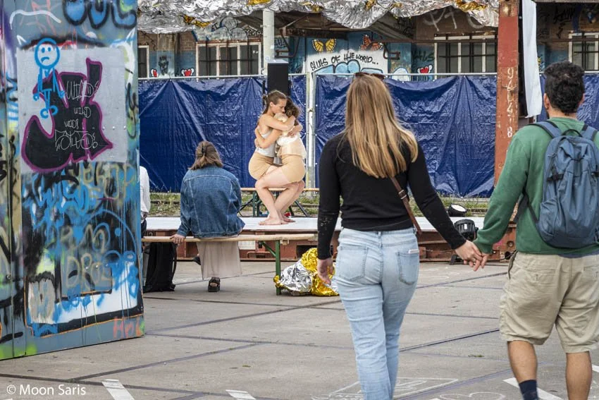 Two people, a man and a woman, walk hand in hand in a parking lot towards a makeshift stage where a woman is performing or preparing to perform, with several people seated nearby. The background and surroundings feature graffiti art and a blue tarp c