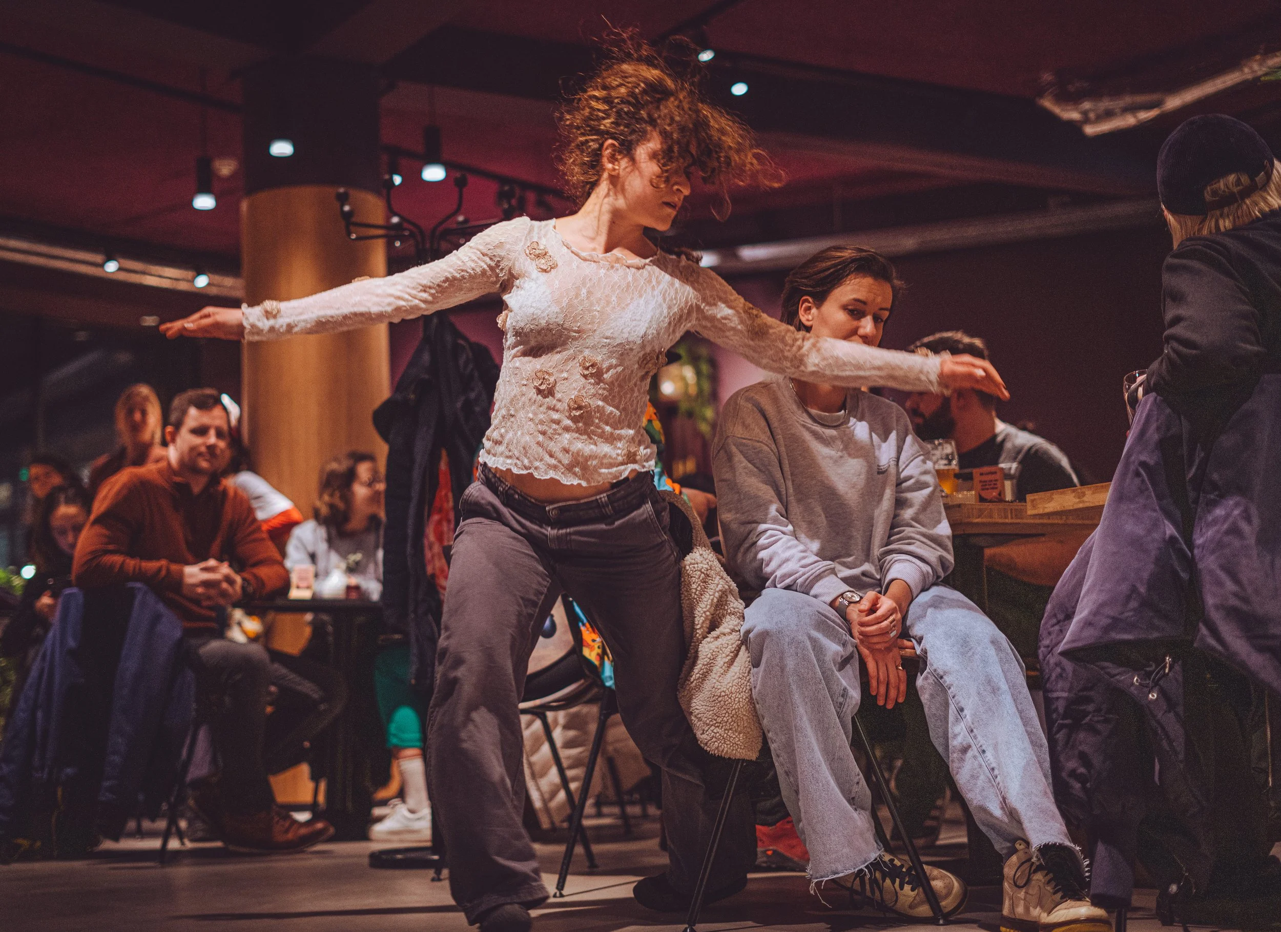 A woman with curly hair is dancing at a social gathering, with her arms extended to the sides. She is wearing a distressed white long-sleeve top and dark jeans. Others are seated around her, including a woman in a gray sweatshirt and light jeans, wat