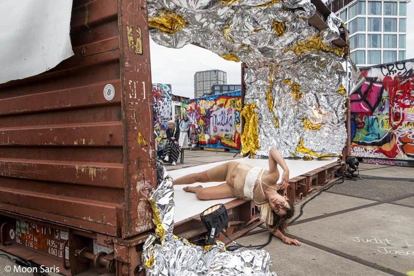A performer lying on the ground next to a makeshift stage in an urban outdoor setting. The stage is constructed from a shipping container with metallic foil and colorful graffiti decorations. Buildings are visible in the background.