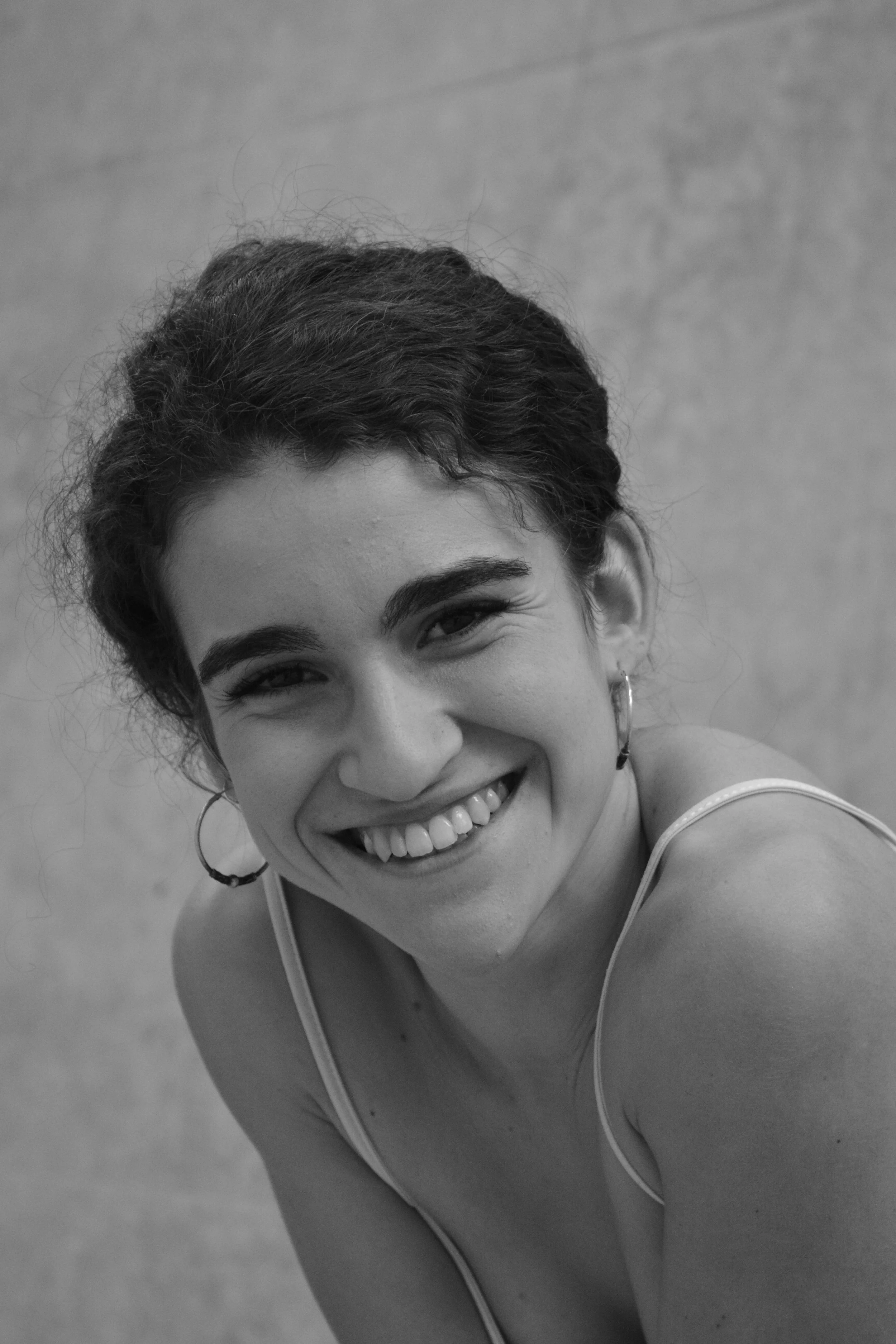 Black and white photograph of a young woman smiling, wearing hoop earrings and a sleeveless top, with a plain wall in the background.