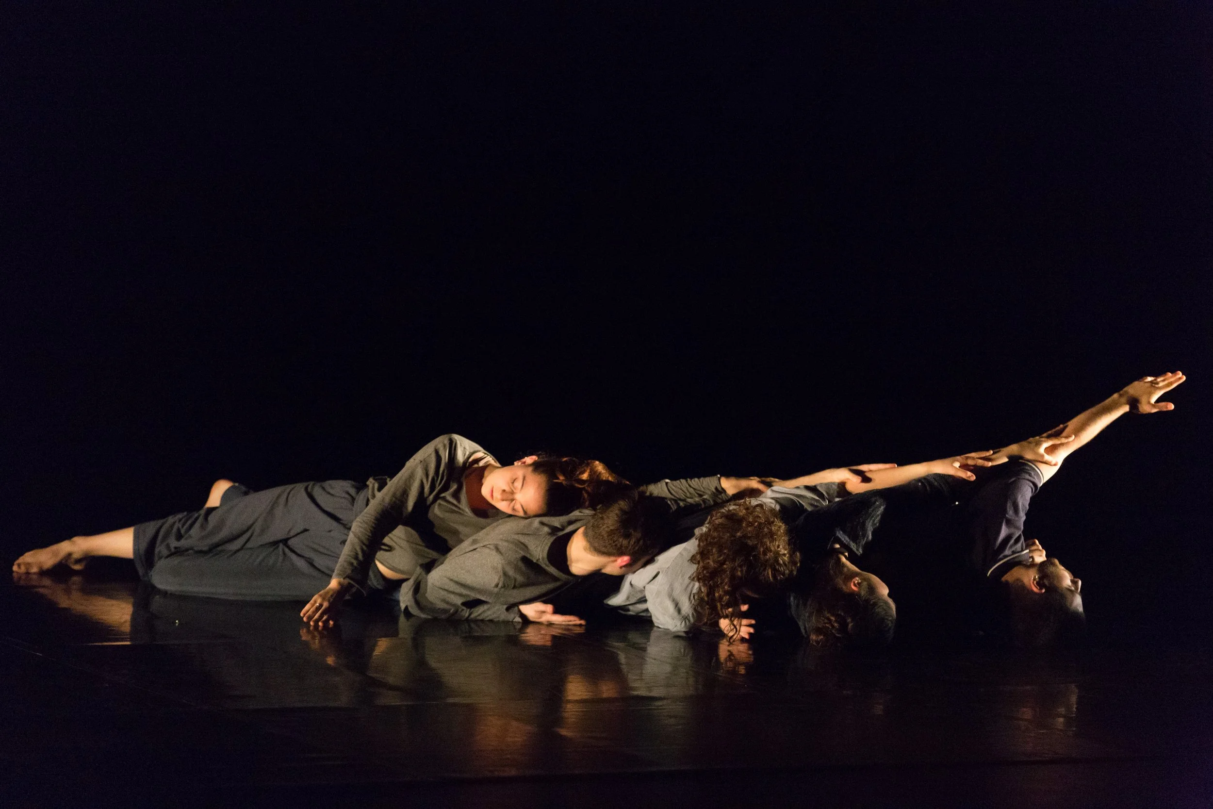 Five dancers performing a contemporary dance on a dark stage, all lying on the floor in a line with arms and legs extended, wearing dark clothing.