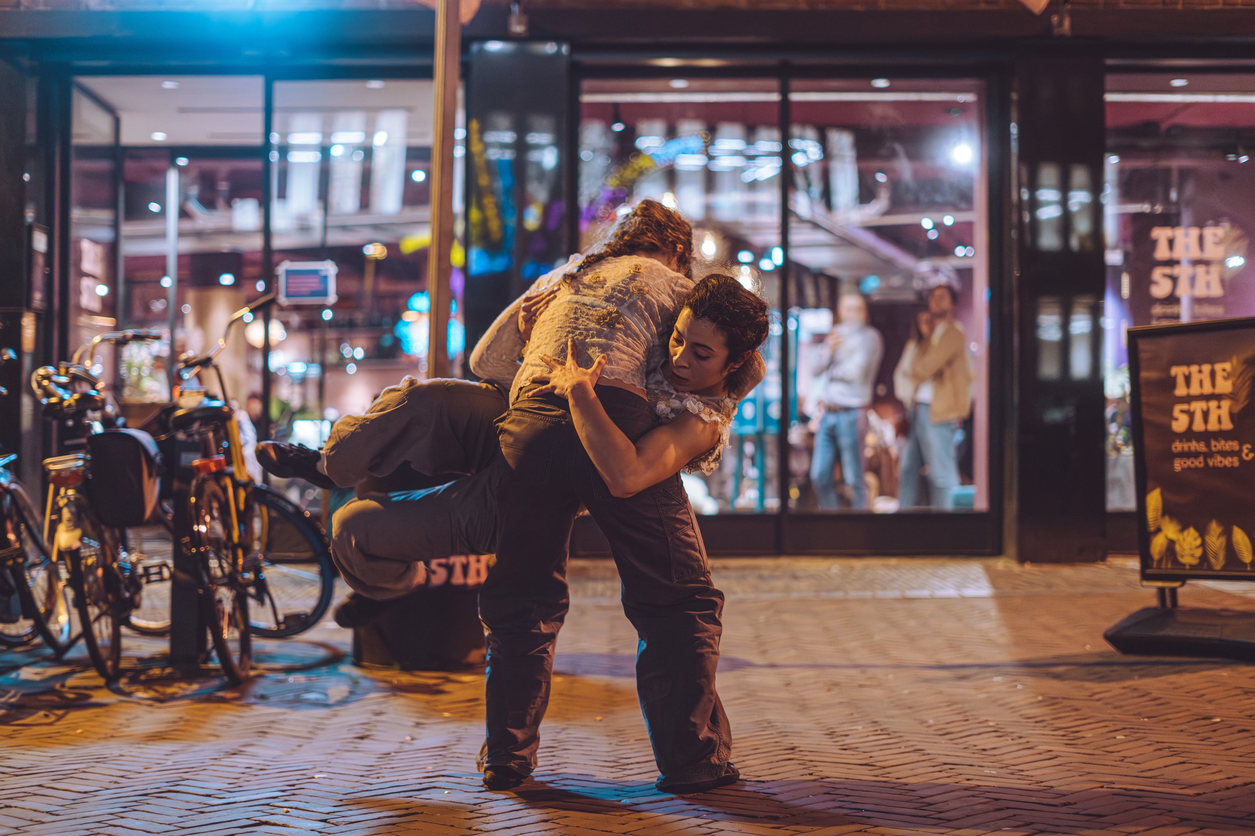 Two women engaged in a physical fight outside a restaurant or bar at night. One woman is holding the other in a headlock, and the woman being held appears to be struggling. There are bicycles parked nearby, and a reflection of people inside the estab